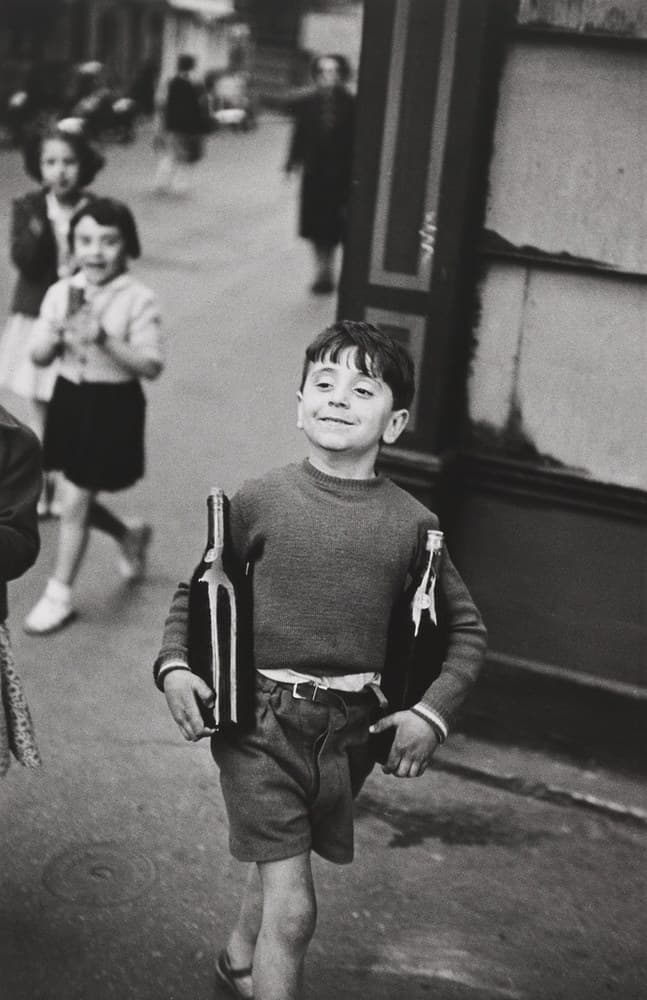 A grinning boy of about seven walks straight towards the camera down a Paris street, a tall dark bottle clamped under each arm with the necks up, his right hand curled under the base of one bottle and his left hand hanging loose and half-closed at his side. He wears a dark ribbed crew-neck jumper with pale-striped cuffs, a pale shirt tucked in under a dark leather belt with a bright rectangular buckle, dark pleated shorts and open sandals, and his head is tipped back. Two girls follow a few paces behind on his right, the taller in a dark jacket with bobbed hair laughing with her mouth open, the smaller in a pale cardigan and dark skirt with white ankle socks and white shoes, caught mid-step with her hands clasped at her chest. A sliver of a fourth figure in spotted fabric is cut by the left edge, and three blurred adults walk further up the street. The right third of the frame is a dark panelled wooden shopfront pilaster beside a cracked, water-stained rendered wall with no lettering on it