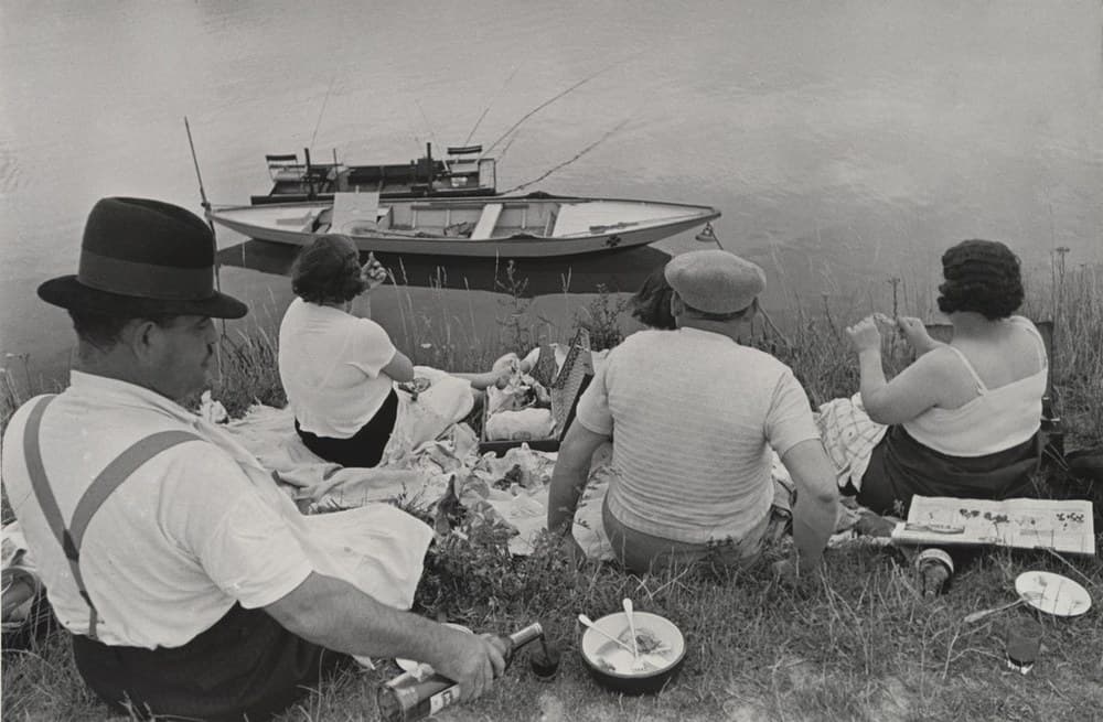 Five people sit on a grass riverbank with their backs to the camera. Nearest, at the left, a man in a dark fedora with a ribbon band and a white short-sleeved shirt, pale braces crossing his back in an X, reaches down to take a bottle by the neck at the bottom edge, his head turned far enough away that only an ear and the tip of his nose show. Behind him a woman with short dark hair in a white top raises one hand in front of her. At the centre a heavier man in a pale peaked flat cap and a ribbed shirt sits braced on one arm, with a fifth dark-haired head just visible past his shoulder, and at the right a woman in a white sleeveless top, her dark hair set in deep waves, holds both hands up in front of her. A white cloth spread with food, a woven basket with its lid propped open, an open newspaper, a dark-rimmed bowl with a spoon and fork in it, a white plate and a fallen bottle lie on the grass around them. Flat pale water fills the whole upper half and runs off the top edge with no far bank and no sky, and a long low wooden boat with a four-lobed motif painted near its bow and two thin fishing rods angled out to the right is moored across the middle