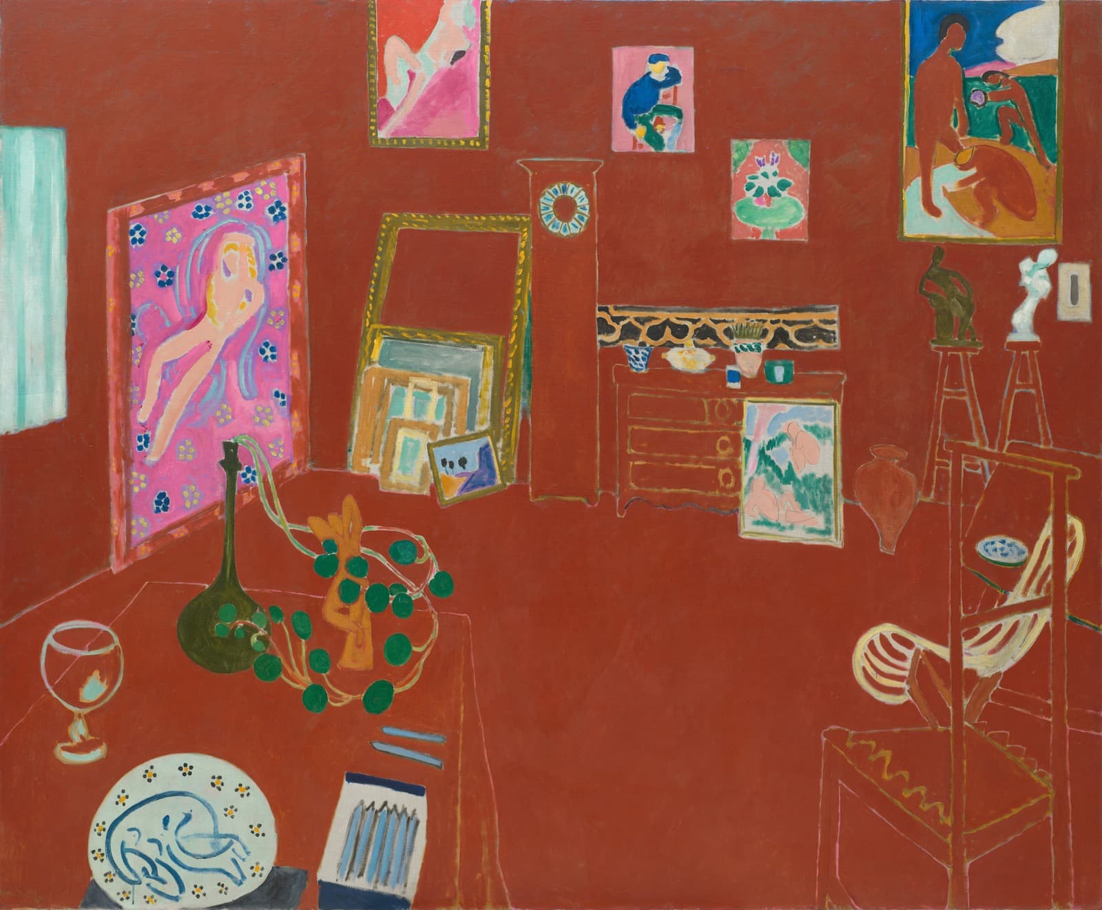 A studio interior painted almost entirely in one brick red. Framed canvases in their own colours hang on the walls. A nude lies on a magenta flowered ground at the left, a small unframed picture of a seated man in a blue smock hangs at the centre, and a terracotta nude stands against a cobalt sky at the upper right. A chest of drawers, a grandfather clock with no hands, a table and two stools appear only as thin pale lines drawn on the red. A ceramic plate, a stemmed glass, a green bottle, a trailing nasturtium and a box of crayons stand on the table in the foreground