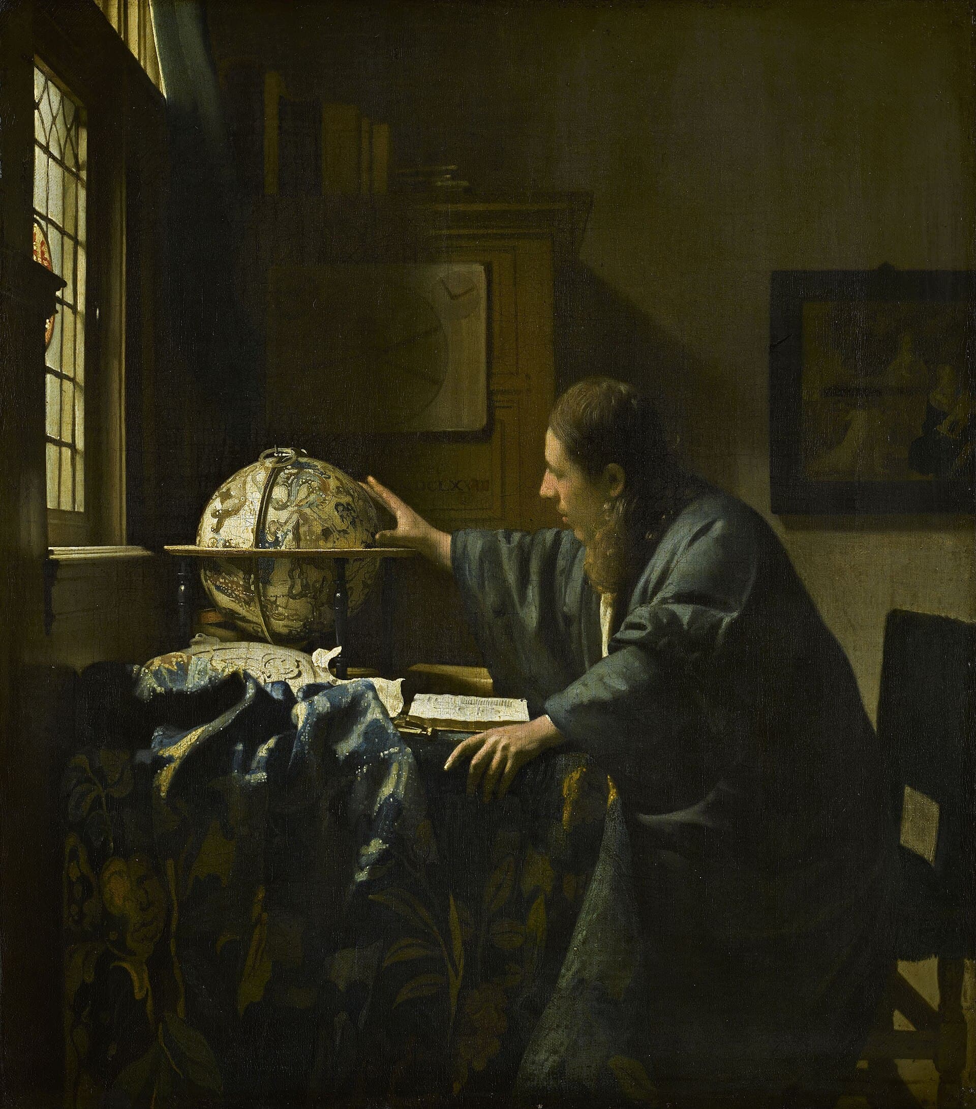 A long-haired man in a loose blue-green robe leans over a table covered with a flowered cloth and reaches toward a globe of the stars. An open book lies in front of him, light comes from a leaded window at the left, and a dark framed painting hangs on the wall behind him