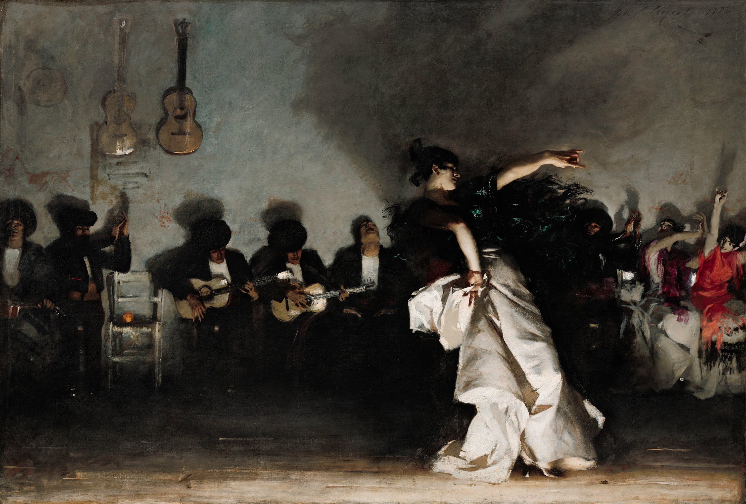 A flamenco dancer leans back in a low gray room, her left arm flung up, a black shawl over her shoulders and a heavy white skirt gathered in her right hand; light from floor level brightens her arms and skirt and leaves her face dark, throwing a large shadow on the wall. To her left a row of seated men in black play guitars, clap and sing; two women in red and magenta clap at the right; two guitars hang on the wall and an orange sits on a white chair.