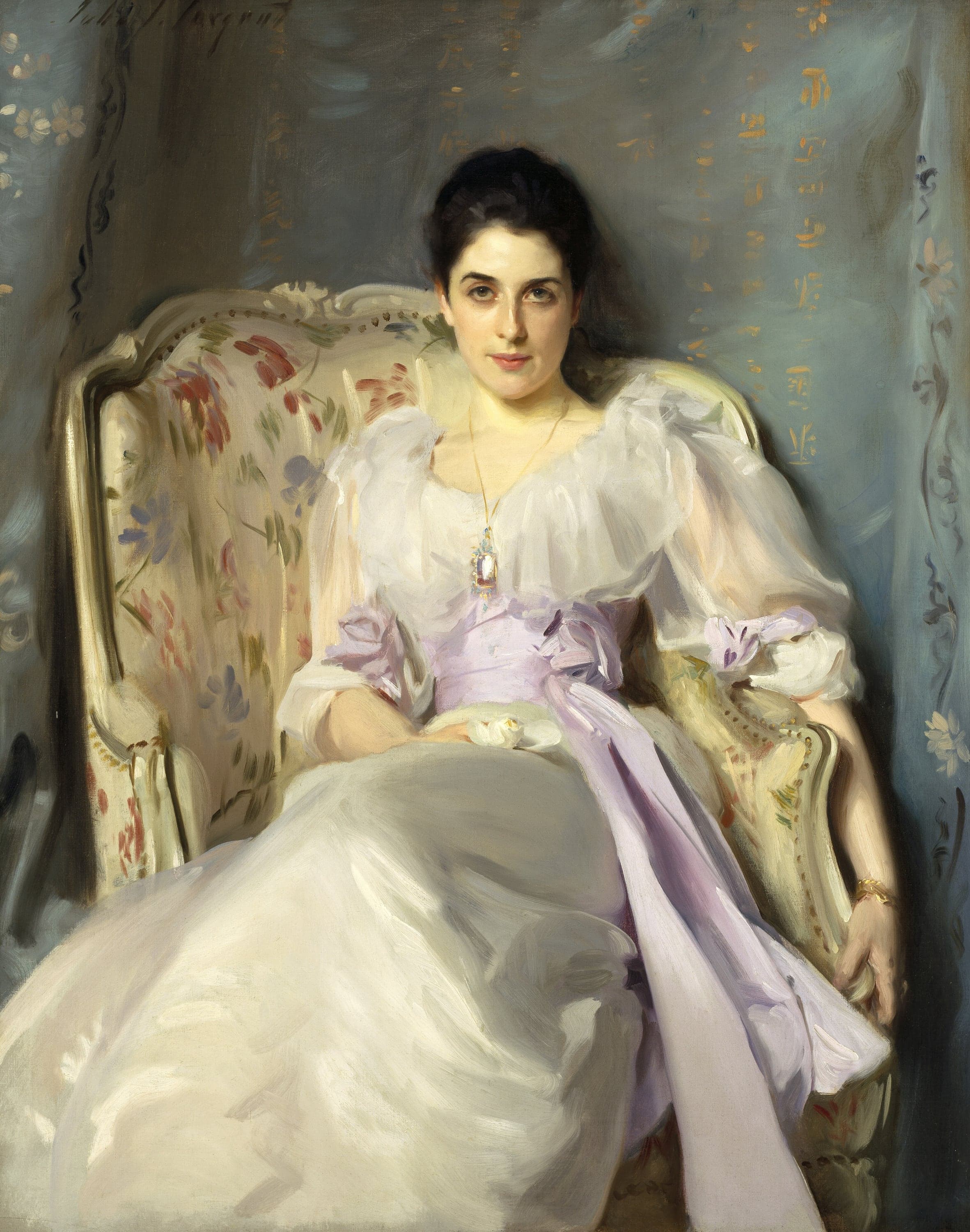 A dark-haired woman in a white dress with full gathered sleeves and a lilac sash sits back in a cream armchair patterned with red and blue flowers, one arm along its arm, a jeweled pendant at her throat, looking directly out at the viewer against a pale blue-green silk hanging.
