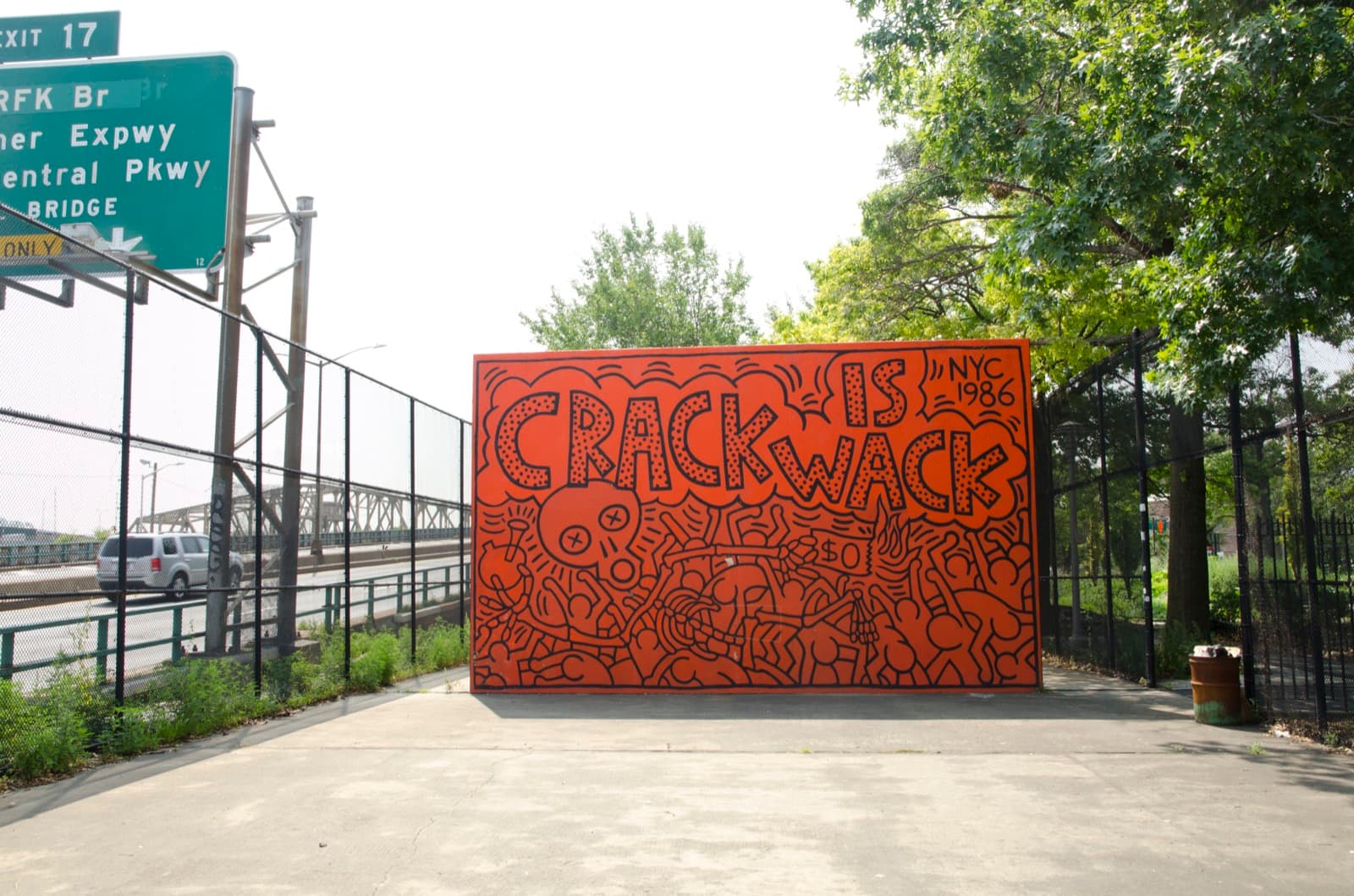 An orange wall standing on a paved court beside a highway, lettered CRACK IS WACK across the top in fat black capitals filled with dots, each word inside a cloud-shaped bubble, with NYC and 1986 in plain characters at the top right. Below the lettering the orange field is packed with black-outlined figures, a large skull with a circled X for each eye, a figure holding a rounded vessel on a stem at the lower left, and a torn dollar bill marked $0 with three tongues of flame rising from it. A green highway sign reading EXIT 17 and RFK Br stands at the upper left, with a chain-link fence, the Harlem River Drive and trees around the court