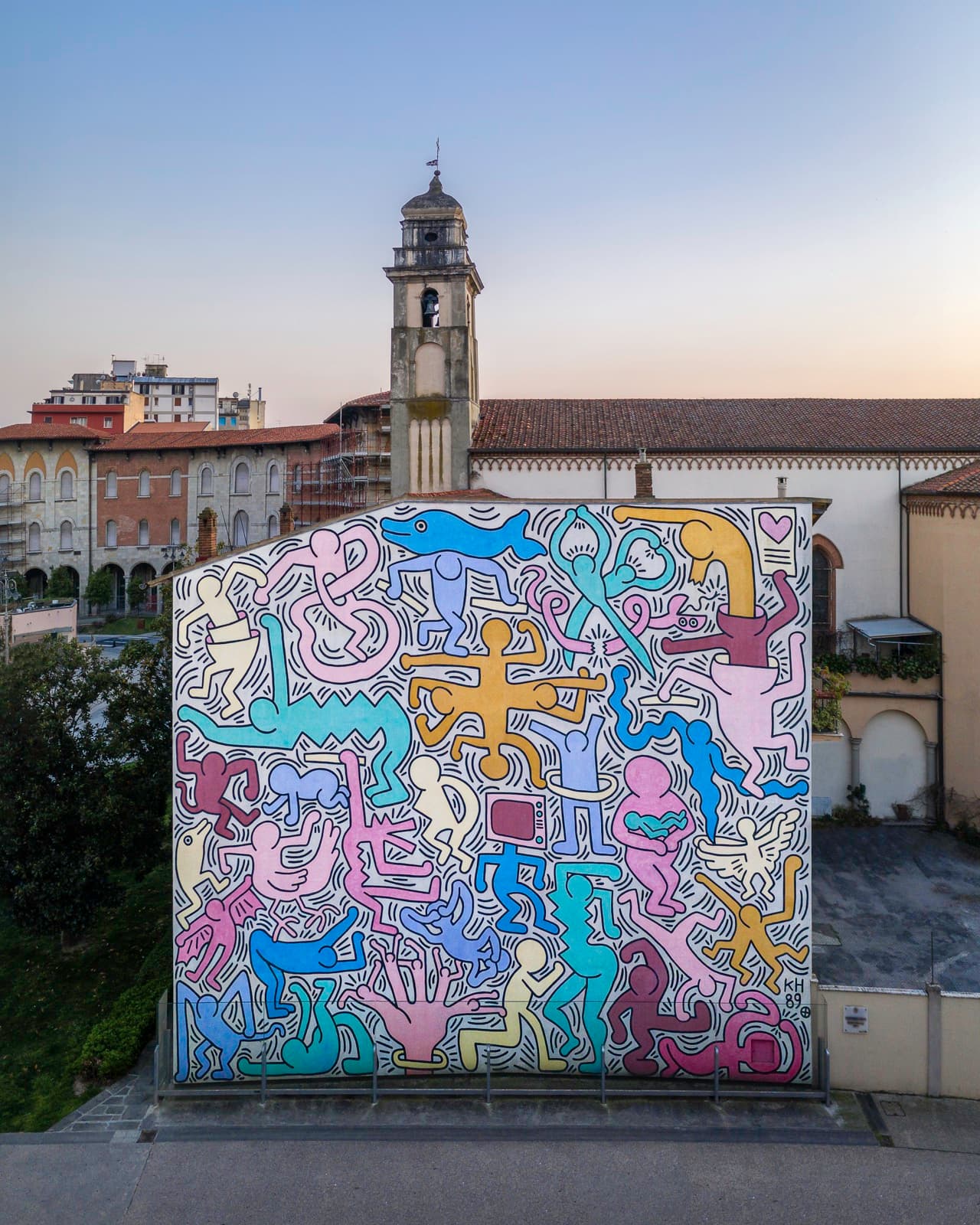 A tall church wall seen from above, filled corner to corner with thirty interlocking figures in heavy black outline coloured in flat pink, lilac, ochre, blue, cream and dark red, with short parallel strokes packed into every space between them. A blue dolphin runs across the top with a lilac figure carrying it on its back, an aqua figure drawn as a pair of open scissors cuts into a coiling pink snake at the upper right, a large pink figure holds a small turquoise baby across both forearms below centre, a television with a dark red screen stands on a blue body, and a cream winged figure is at the right. The initials KH and 89 are painted at the lower right. The church's stone bell tower and tiled roof rise behind the wall, with Pisa's render-fronted buildings on either side