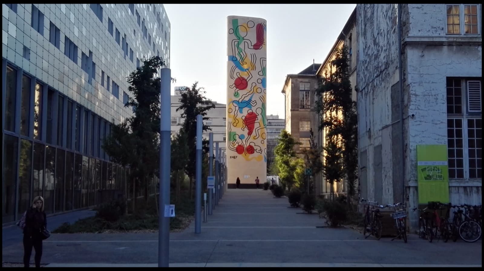 A tall narrow concrete stair tower standing at the end of a paved way between hospital buildings, painted from top to bottom on a pale cream ground with black-outlined figures over flat patches of red, yellow, blue and green. A striding figure with a plain round head holds the top, tumbling bodies fall down the shaft past red discs, and a figure on all fours sits near the foot. The initials KH87 are painted at the lower left of the painted area. A glass-fronted building runs down the left of the way and older stone buildings down the right, with two people standing at the base of the tower