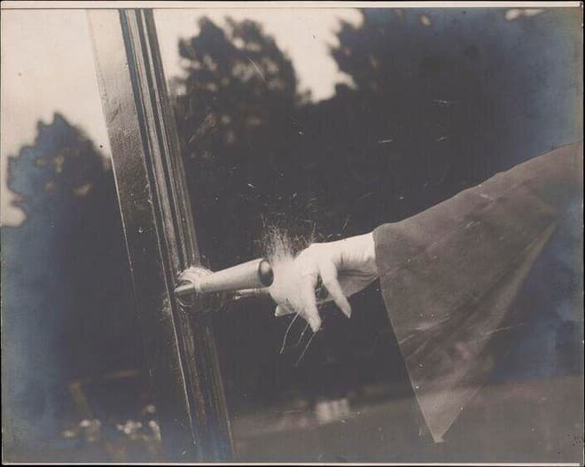 A black and white photograph of an arm reaching in from the right in a dark coat with a wide cuff. The pale hand is open with its fingers hanging, a dark bangle at the wrist, and a spray of fine white threads bursting upward from behind the bangle across dark glass. The arm runs left to a metal lever handle on the upright of a glass door, whose moulded frame crosses the left of the picture. Trees and a patch of bright sky are reflected in the glass