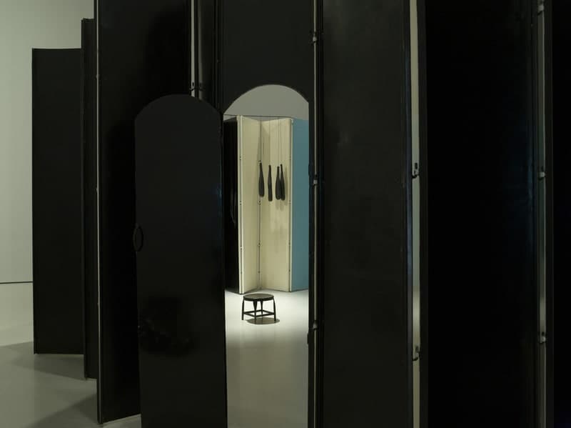 Tall matt black steel panels stand edge to edge across a pale gallery floor, hinged together with visible vertical pins, some with rounded tops and some square. A gap between two of them opens onto a small white-floored room inside. Within it a screen of cream-painted panels folds around a corner beside one flat pale blue section, and four black rubber shapes hang on thin threads against the cream, each narrow at the top and swelling to a heavy rounded end. A small dark stool with a round seat and four splayed legs stands alone on the white floor, casting one shadow