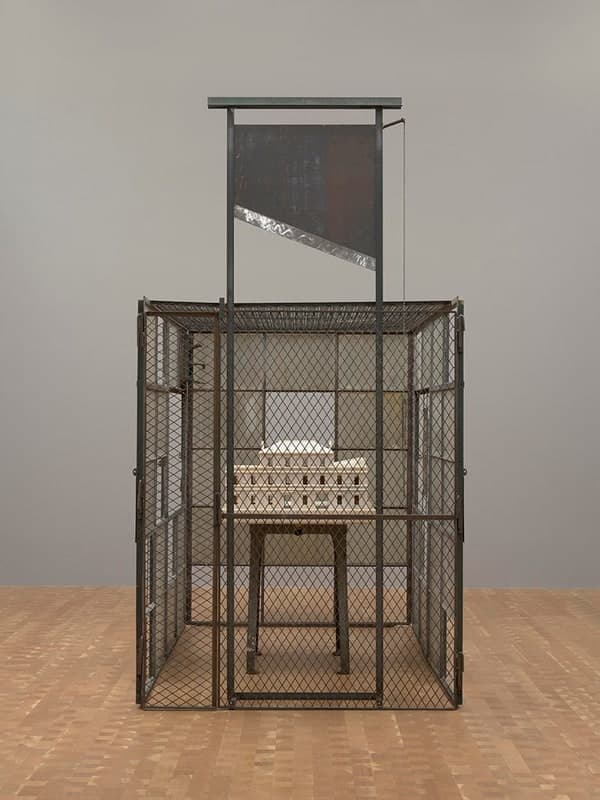 A tall rectangular cage of diamond wire mesh in a rusted metal frame, standing on a pale parquet floor against a plain grey gallery wall and photographed square on. Inside the cage a long white marble model of a house with a pitched roof and three ranks of small window openings rests on a plain four-legged table. Above the cage a separate steel frame holds an upright dark rusted panel with a bright silver blade set diagonally across it and a thin chain hanging down the right-hand post. A hinged door with a heavy latch is visible at the left front corner