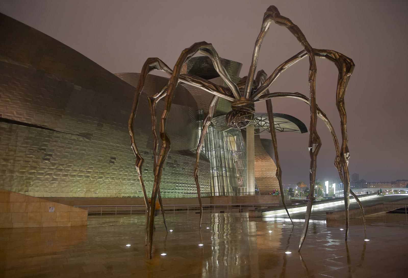 A bronze spider about nine metres tall standing at night on wet paving outside a museum. Eight ribbed legs rise to high pointed apexes and taper down to thin tips, each set in a shallow puddle lit by small round floor lights. The body hangs high between them: a ribbed, coiled abdomen above a collar, and below it an open basket of wire mesh studded with small bulges. Behind the spider are the titanium-clad curved walls of the Guggenheim Museum Bilbao and a tall glass atrium, with a lit walkway, palm trees and city buildings to the right under a low brown night sky