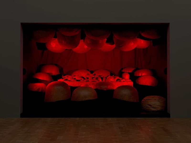 A dark chamber set into a grey gallery wall above a parquet floor, seen square on and lit entirely in red. A close row of rounded soft forms hangs from the ceiling, each glowing red underneath. A second row of matching domes sits along the floor at the front, lit dimly so they read dull brown. Across the middle a low platform carries a heap of small indistinct shapes lit bright red. Loose folds of fabric hang at the back, and one pale unlit lump sits apart at the lower right