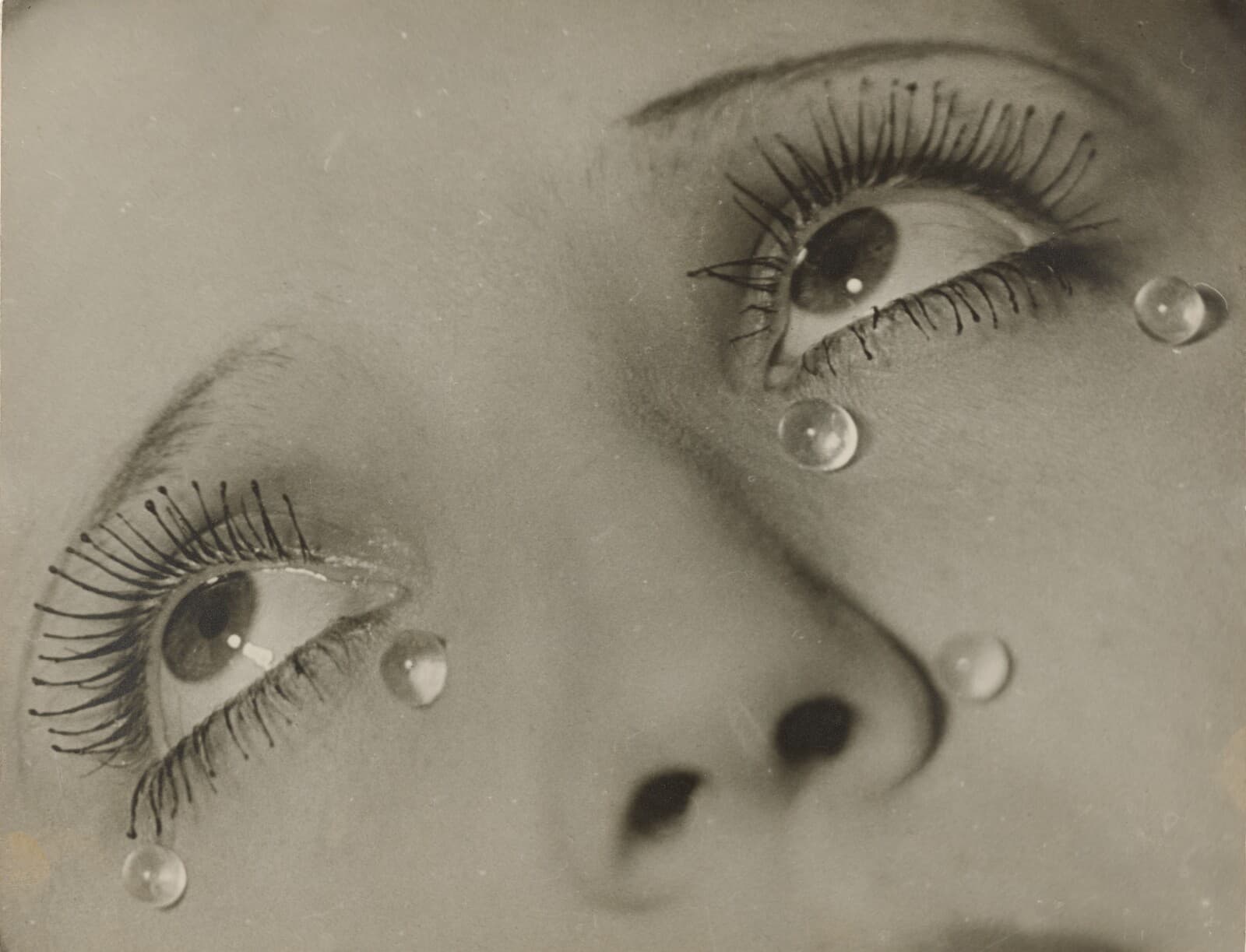 A close view of the upper half of a woman's face, cut at the brows and below the nostrils, the head tilted back and both eyes turned up and away to the right. The lashes are heavily mascaraed, standing apart as stiff spikes with rounded black tips on one side and finer and doubled on the other. Five clear glass spheres rest on the skin below the eyes, one of them hanging from a lower lash, each holding a bright point of reflected light