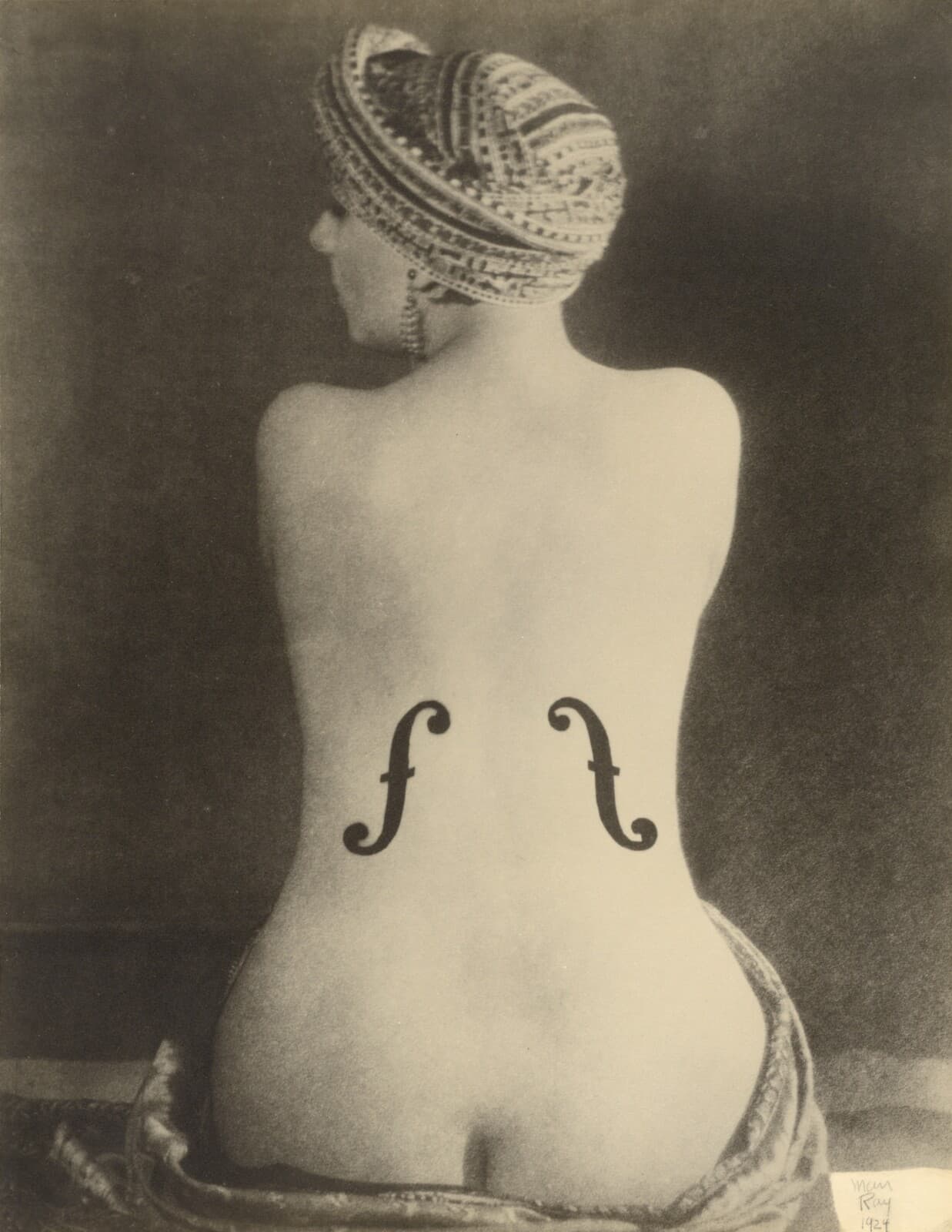 A woman sits with her back to the camera, bare to the hips, her head wrapped in a patterned turban and turned so that only the edge of her profile shows. A long beaded earring hangs clear of her jaw. Two solid black violin f-holes, each with a round eye at either end and a short nick at mid-height, stand on her lower back on either side of the spine, the right one set higher than the left. Patterned cloth is bunched under her hips, and the backdrop darkens from left to right
