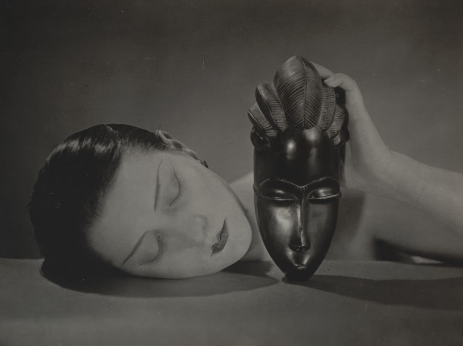 A woman's pale face lies sideways on a table, eyes closed, her short dark hair slicked flat. Beside it a black polished wooden mask with a long oval face and half-closed eyes stands upright on its chin, held at the top by her hand, its crest carved like grooved leaves. Both cast soft shadows on the tabletop against a plain grey background