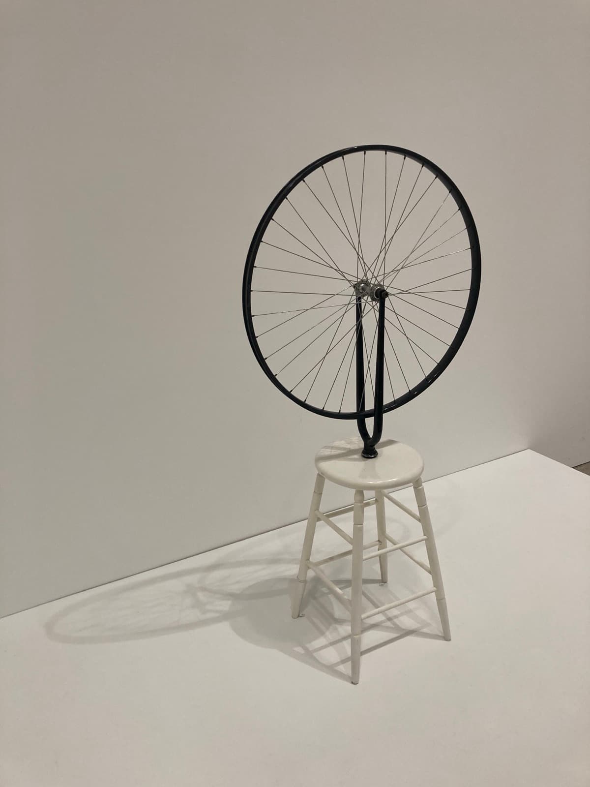 A bicycle wheel with no tyre, mounted upside down on a white-painted wooden stool in a gallery. The black fork is inverted so the wheel rides at the top and the fork's neck runs down into the centre of the round stool seat. The wheel's bright metal spokes cross between a bare metal hub and a black-painted rim. The stool has four splayed turned legs joined by two tiers of stretchers. The wheel casts a long flattened shadow across the white plinth to the left