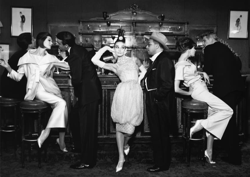 Eight people along a carved bar with a tall mirrored back fitting and rows of stemmed glasses. A woman with a high topknot stands at the centre in a beaded dress with a jewelled collar, bubble hem and long white gloves, one hand raised to her hair. A man beside her wears a dark tuxedo, a pale wide-brimmed hat, spectacles and a lit cigar. At the left a woman perches on a stool in a pale suit with an enormous stand-away collar while a man leans towards her; at the right a woman arches backwards over a stool as a man leans over her; a barman in a bow tie stands behind the counter