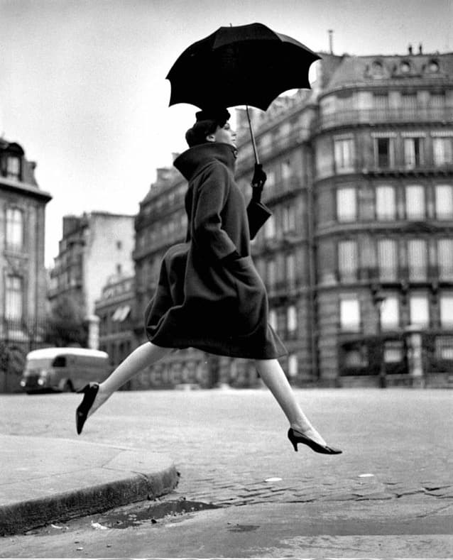 A woman in a full dark coat is caught in mid-air above a wet Paris street, both feet off the ground, the trailing foot pointed behind her. She holds a large black umbrella open and tilted back above her head, wears a small dark cap set back on her hair and dark gloves, and a dark angular bag hangs from the hand gripping the umbrella. Out-of-focus Haussmann buildings and a pale parked van stand behind her under a blank white sky, with a kerb and a puddle in the foreground