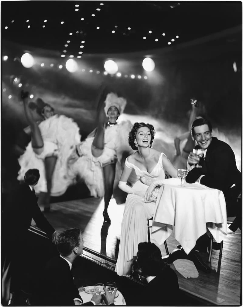 A woman in a pale off-the-shoulder evening gown of fine pleated bands sits at a small round nightclub table with a white cloth, laughing with her head tipped back, long gloves ruched down her forearms and her gloved hands folded in her lap. A man in a dark dinner jacket beside her raises a stemmed glass towards the camera. Behind them two chorus dancers in white ruffled costumes kick high and are blurred by movement, under a dark ceiling scattered with small lights and a row of large globe lamps, with two seated spectators in the foreground seen from behind
