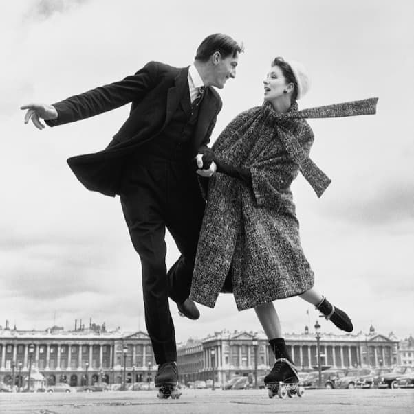 A man in a dark three-piece suit and a woman in a full flecked tweed coat skate across a wide paved square on four-wheel roller skates, hands joined, laughing at each other. He leans steeply back with one arm flung out and one skate lifted; she wears a pale hat set far back, dark gloves and dark ankle socks, and a long tweed stole streams out horizontally behind her. Two matching colonnaded palace fronts, a tall ornate lamp standard and a row of parked 1950s cars stand behind them under a flat pale sky