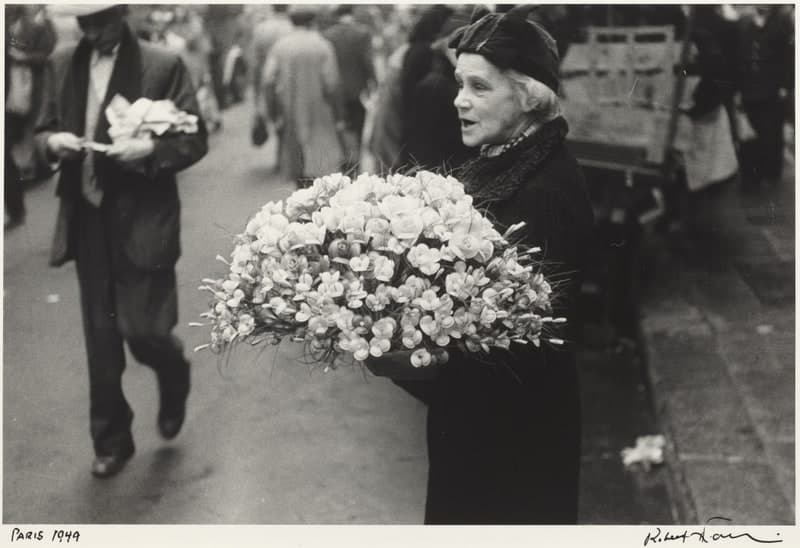 A black-and-white street photograph. An elderly woman in a dark coat, a heavy knitted collar and a dark cloth cap over white hair walks from right to left with her mouth open, carrying a wide armful of small pale flowers on long stems across her front. Behind her a man in a flat cap and dark suit walks toward the camera holding a paper-wrapped bunch, out of focus, with a blurred crowd behind him. A wooden handcart with slatted sides stands at the right, and two or three loose blooms lie on the paving at the lower right. In the white margin below the image the artist has written Paris 1949 at the left and signed at the right