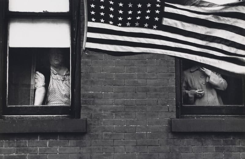 A horizontal black-and-white photograph of a dark brick wall with pale mortar, holding two windows with projecting sills. At the left window a heavy-set woman in a short-sleeved dress with a small repeating print leans forward on the sill, one bare forearm on the ledge, her face behind the glass of the raised sash and under a white roller blind, blurred and shadowed. At the right window a person in a light buttoned coat with a rounded collar stands with one hand raised near chin height and the other at the waist, the head completely hidden by a large American flag that hangs across the upper right of the wall, its canton at the upper left and its stripes billowing off the right edge of the frame
