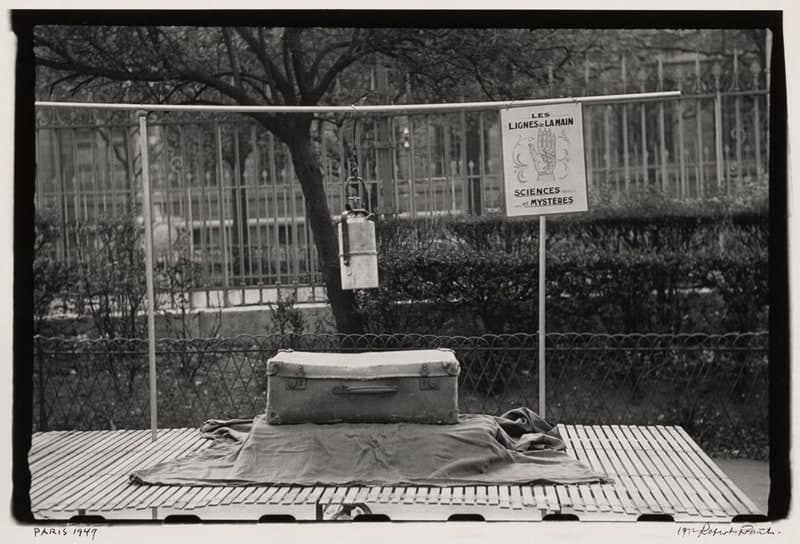 A black-and-white photograph with a printed black border. A flat handcart of narrow wooden slats fills the lower third of the frame, a folded canvas sheet spread over it and rucked up at the right, with a pale battered suitcase standing in the middle, its corners capped in metal. Two thin poles rise from the cart carrying a horizontal bar; a pale metal canister with a side handle hangs from a hook at its centre, and at the right a hand-lettered placard hangs from the same bar reading LES LIGNES DE LA MAIN above a drawing of an open palm and SCIENCES ET MYSTÈRES below. Behind stand a low looped wire fence, a clipped hedge, tall park railings and bare trees. In the margin below, PARIS 1949 is written at the left and the print is signed 1972 Robert Frank at the right