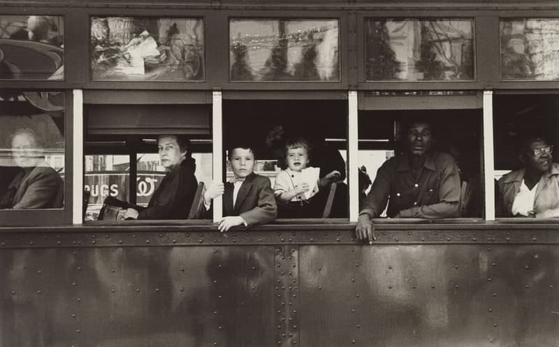 A horizontal black-and-white photograph of the side of a streetcar, cut off at both ends. A riveted metal flank fills the lower third. Above it, five windows hold six passengers in a row: a man behind closed glass at the left, an older white woman in a dark coat looking straight out under arched eyebrows, a white boy in a jacket gripping a white window post, a small child with an open mouth holding a folded white paper, a Black man in a long-sleeved button-down shirt leaning on the ledge on both forearms, and at the right edge a Black woman in glasses looking back over her shoulder. A row of smaller panes above them reflects the street behind the camera, and a shop sign reading DRUGS shows through the first window