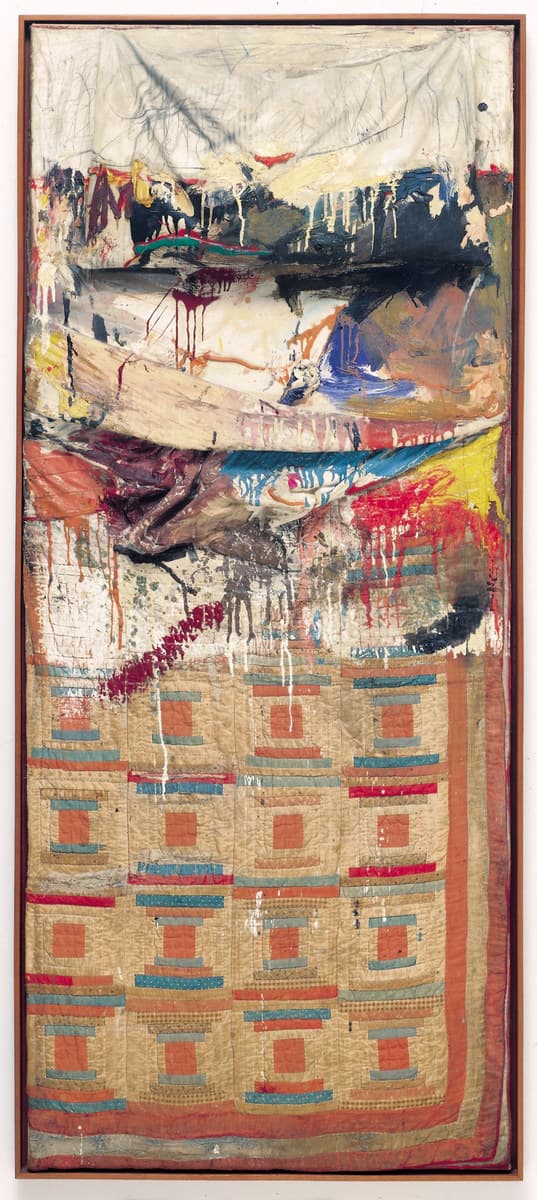 A tall narrow object in a plain reddish-brown frame, hung upright on a pale wall. The lower half is a patchwork quilt in the Log Cabin pattern, each block built around a small orange-red square with bands of blue-grey, red and tan stepping outward, four blocks across, with a rust-orange border down the right edge. Across the middle lies a long pale pillow in striped ticking. Above it a white sheet is pulled taut and covered in dense grey pencil scribbling, and between the pillow and the sheet the surface is loaded with crimson drips, black masses, blue, orange and yellow paint, with cream ribbons of colour running down over the quilt