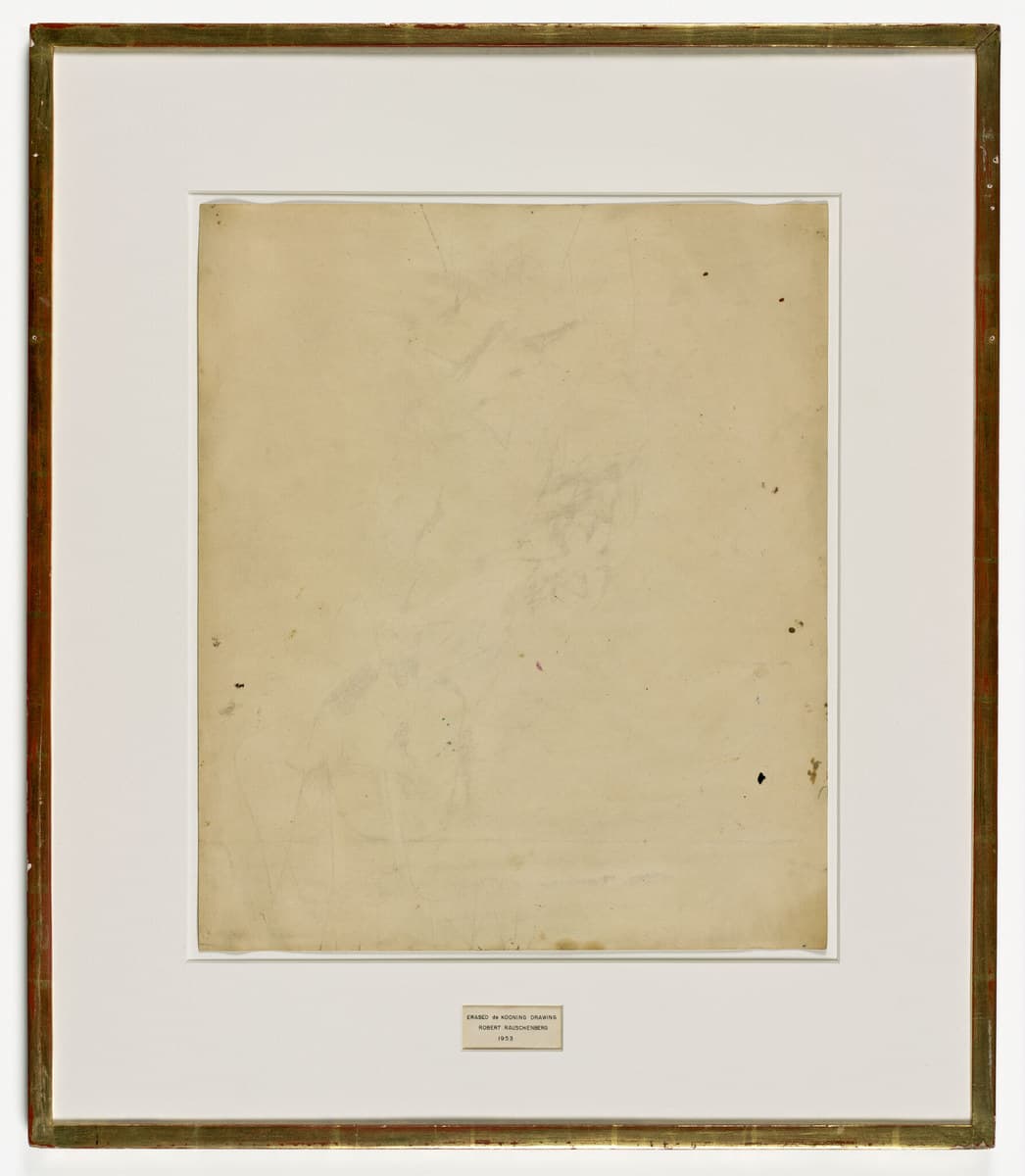 A sheet of cream paper, aged to tan, mounted behind a white mat in a gilded wooden frame whose gilding is worn through to a red ground in places. The sheet looks almost blank, carrying only faint grey traces of rubbed-out drawing, with looping marks and a soft smudge across the upper half, longer strokes at the lower left and a few small dark foxing spots near the right edge. A small cream label in a thin ruled border is set into the mat below the sheet, printed in three lines of capitals reading ERASED de KOONING DRAWING, ROBERT RAUSCHENBERG, 1953