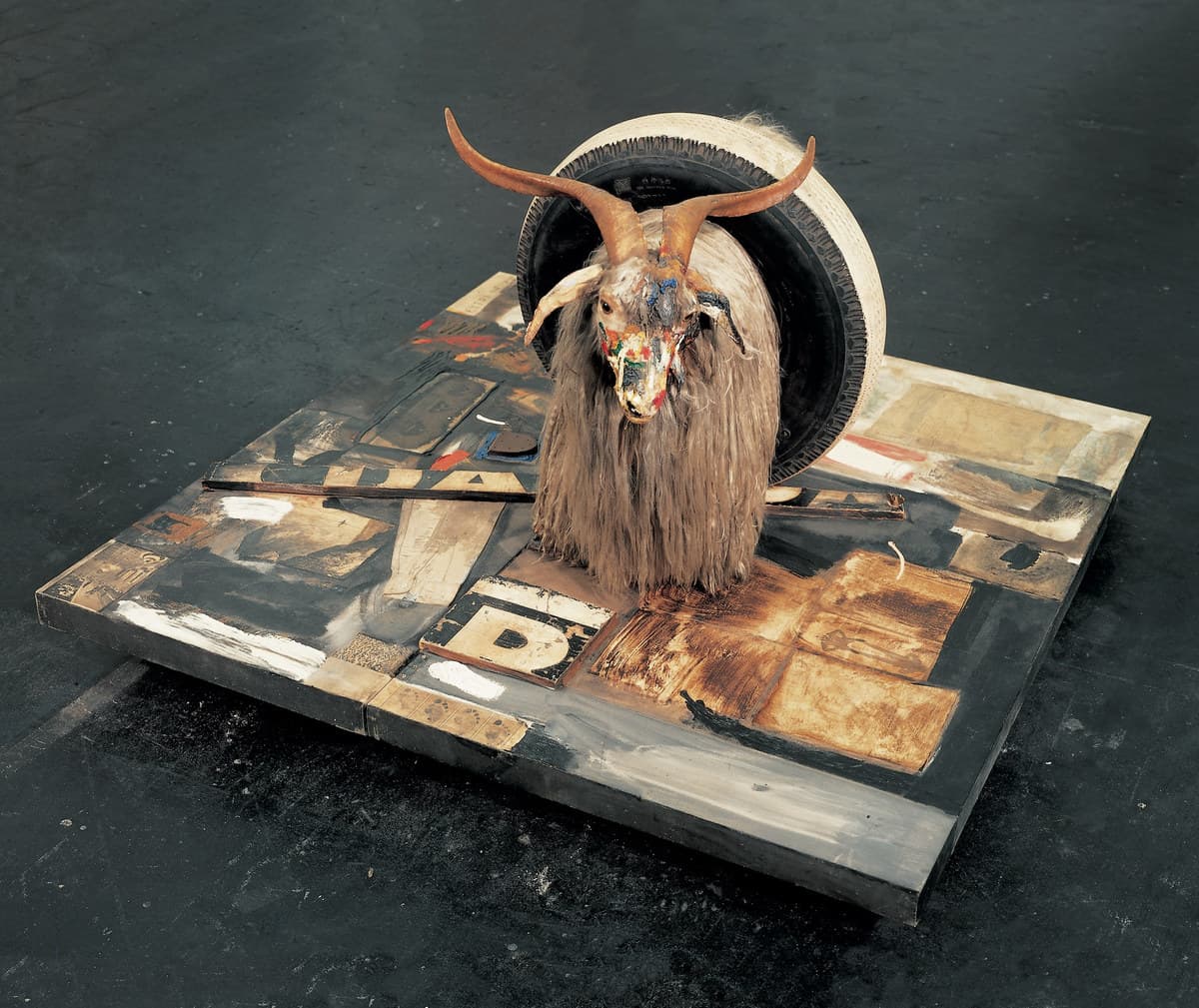 A stuffed long-haired goat with pale curving horns stands facing forward in the middle of a square painted canvas laid flat on a dark concrete floor. Its muzzle is smeared with red, yellow, blue and green paint, and a car tire with white-painted sidewalls is pushed down around its body behind the shoulders. The canvas is worked in greys, blacks and whites with collaged panels set into it, including a board carrying a large black letter D in front of the goat, a wooden lath standing proud across the left half, a rust-brown scorched rectangle behind the forelegs, and a brown rubber shoe heel on a patch of blue paint near the left edge