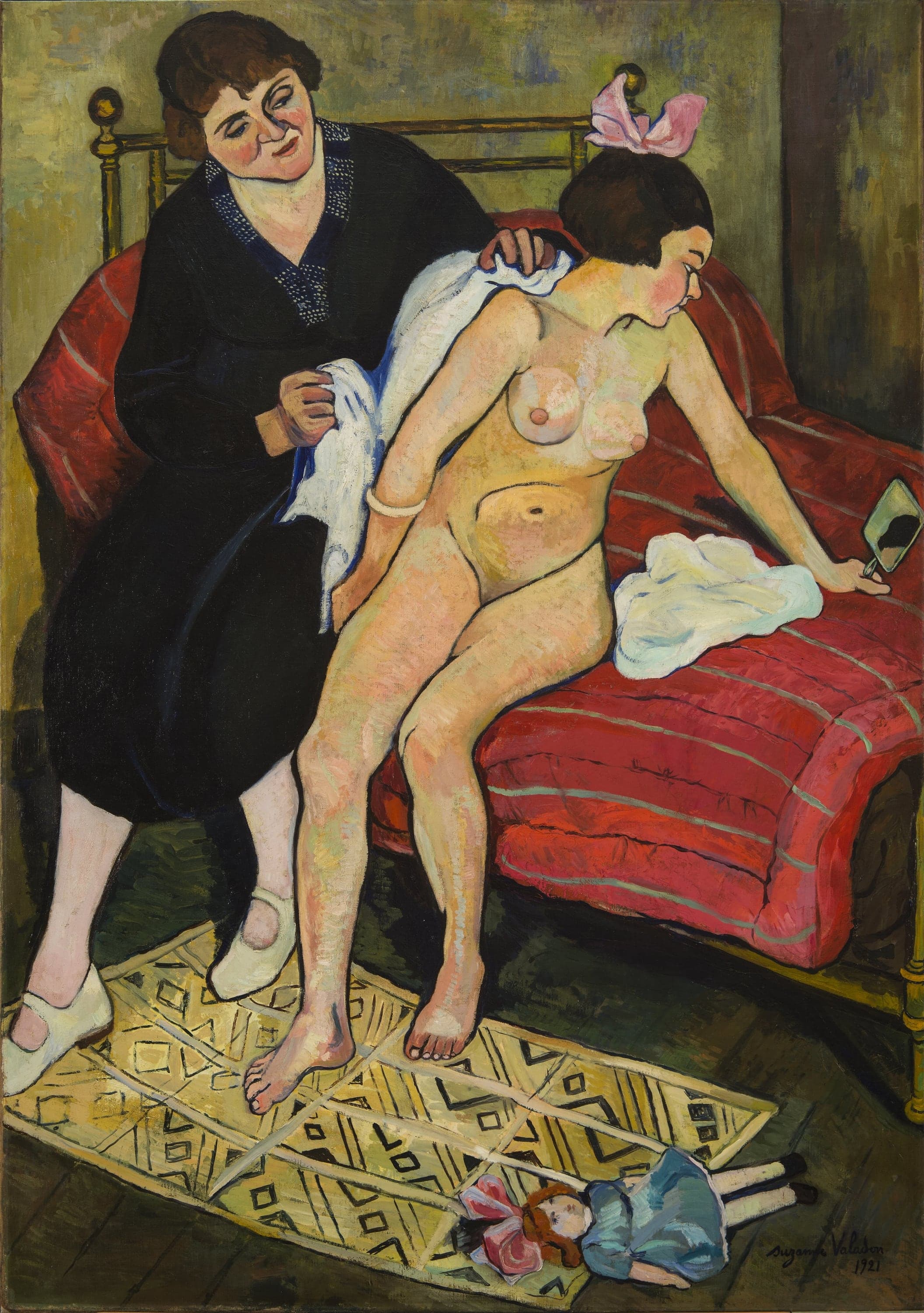 A woman in a black dress with a navy dotted collar sits at the left on a red striped bed, holding a white blue-bordered towel against the back of a naked girl. The girl, a large pink bow on her dark hair and a pale bangle on one wrist, turns away and holds a small hand mirror at the end of her outstretched arm. A doll in a blue dress with ginger hair and a pink bow lies on a yellow patterned rug at the lower right.