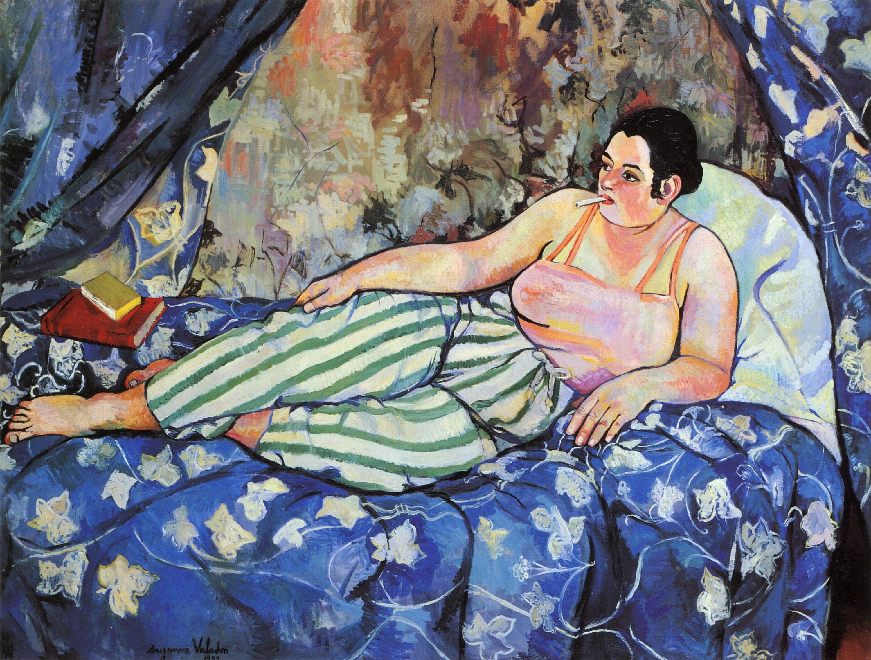 A dark-haired woman lies across a bed, leaning back against a pale pillow, a white cigarette in her mouth. She wears a pale pink camisole with thin coral straps and loose trousers striped green and cream. Two books, a red one lying flat with a yellow one stacked on top, sit behind her single bare foot on a deep blue bedspread patterned with pale cream flowers. A curtain in the same pattern hangs at each side, and a warm patterned wall fills the space between them.