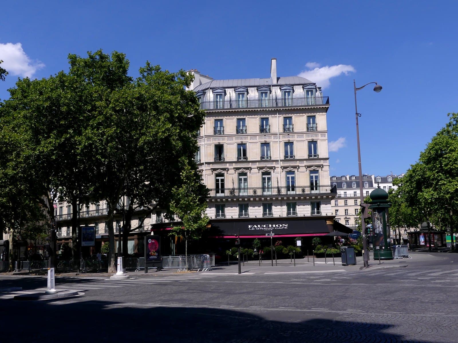 Photograph of the pale stone corner building of Fauchon L'Hôtel Paris on place de la Madeleine, with a dark awning lettered Fauchon Le Grand Café over a planted terrace and trees in front.