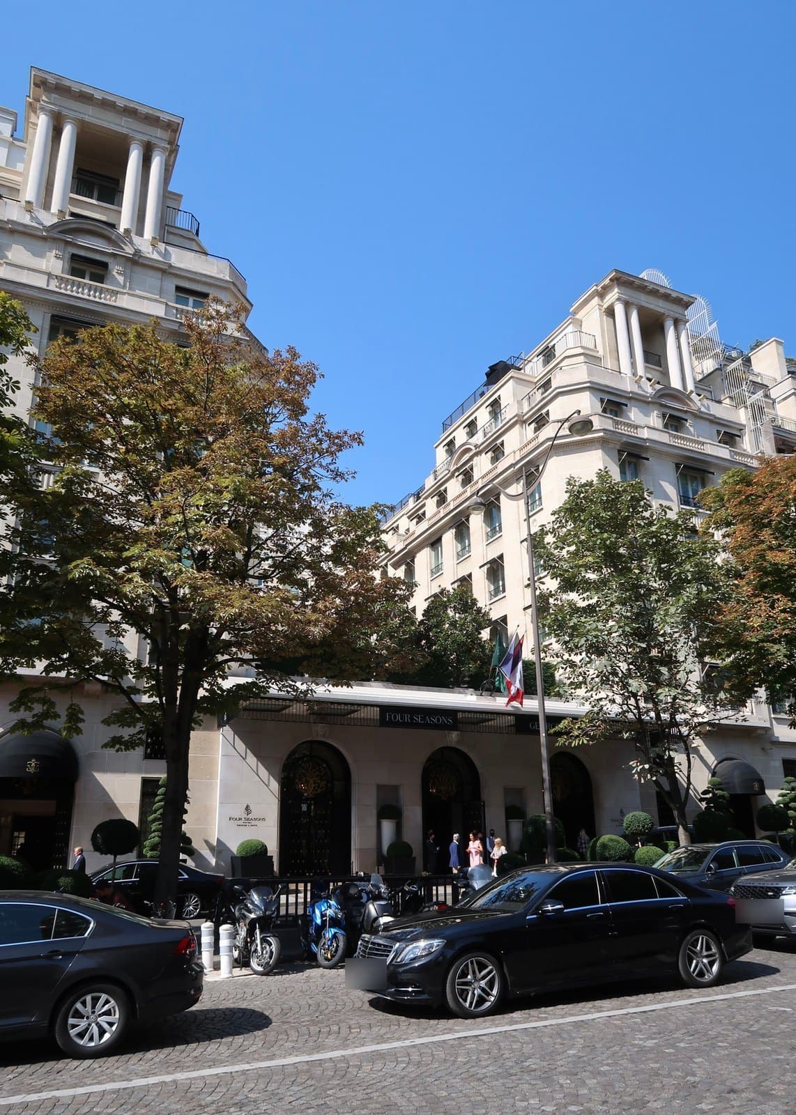 Photograph of the pale stone Four Seasons Hotel George V rising in setbacks to a colonnaded corner tower, with trees in front, a glass canopy over arched doorways, topiary in tubs, a French flag, and black cars and motorbikes parked on the cobbled avenue.