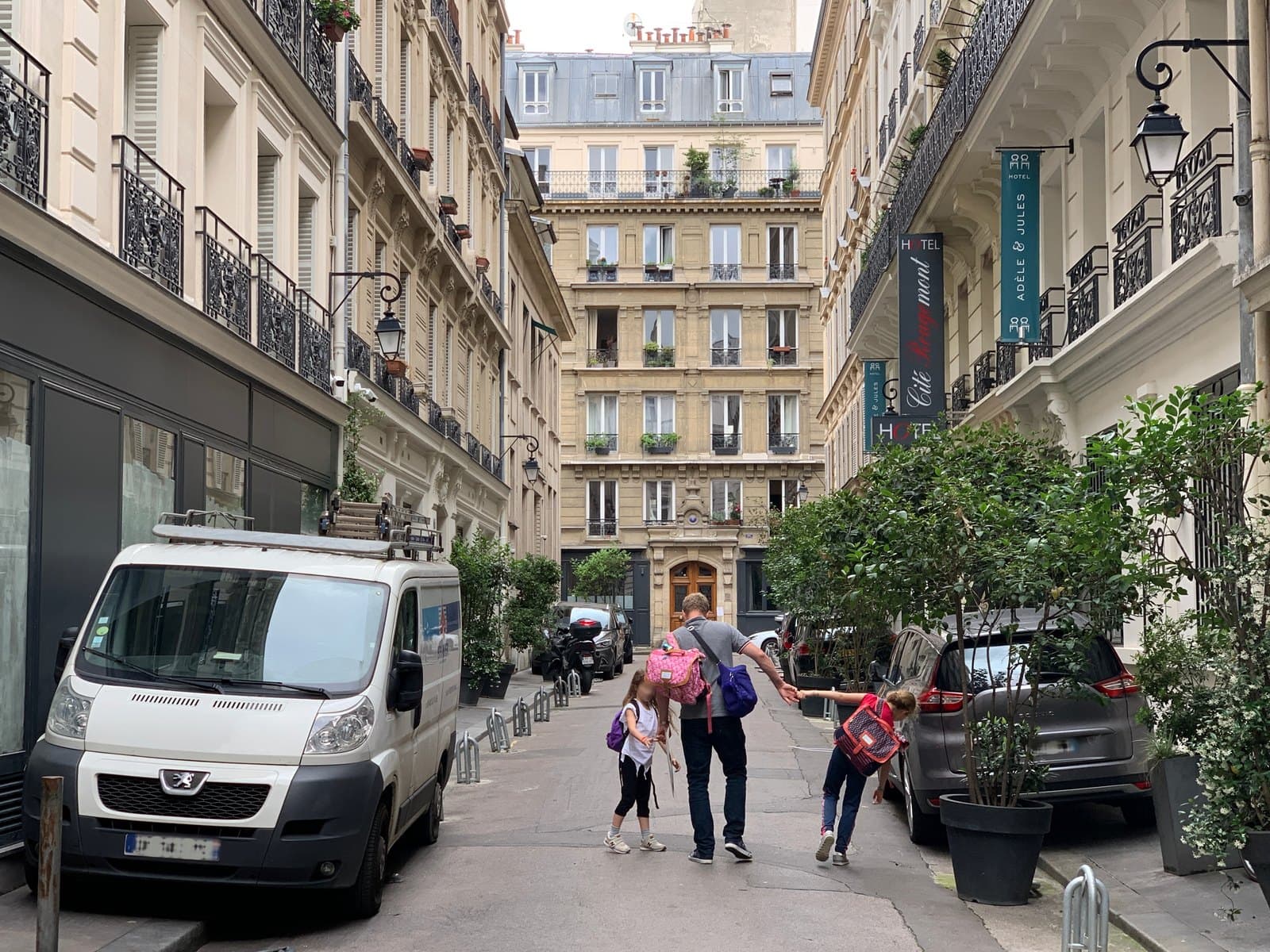 Photograph looking down cité Rougemont, a narrow cobbled lane lined with pale stone buildings, with teal banners reading Hotel Adèle & Jules on the right and an adult walking away with two children.