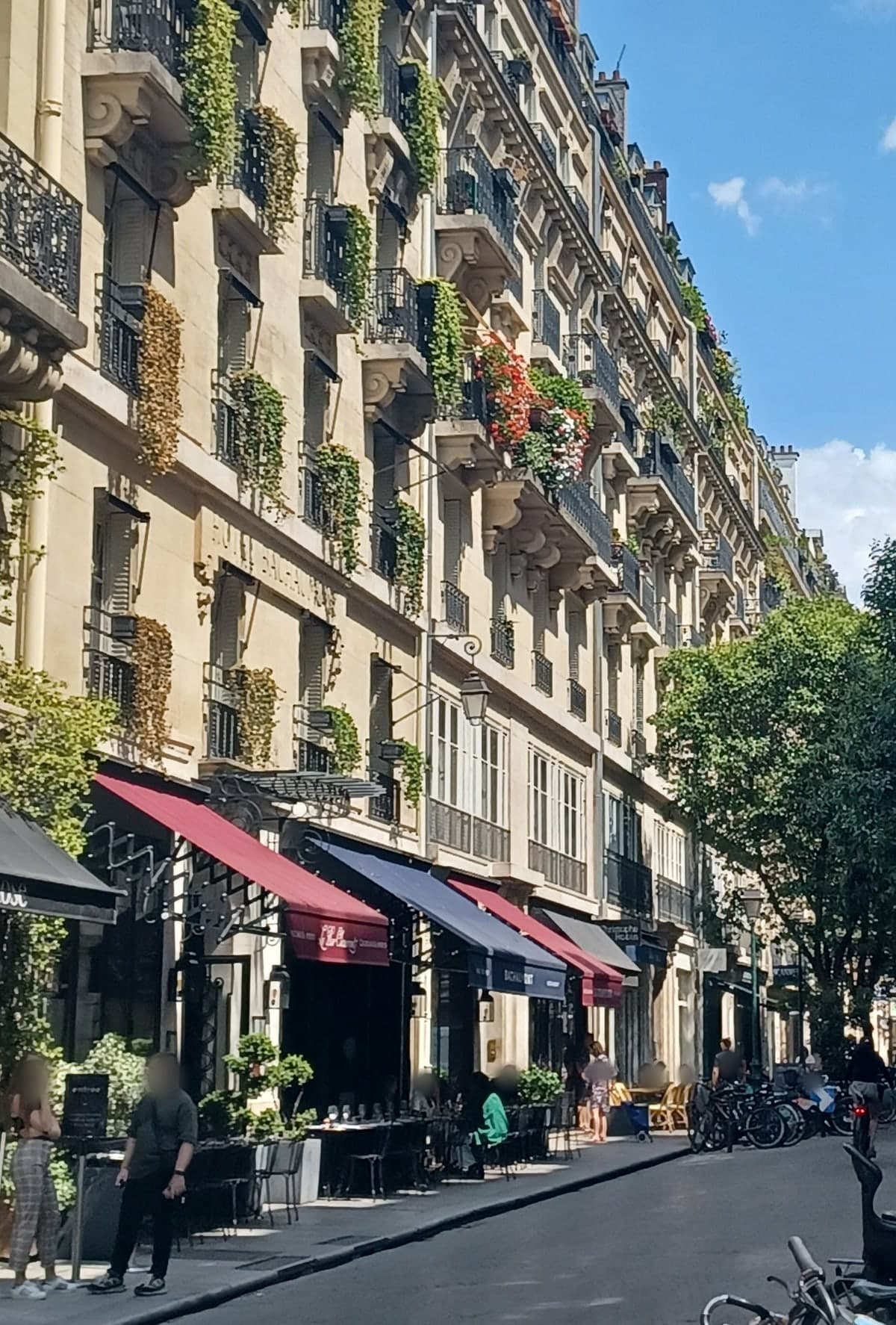 Photograph of rue Bachaumont on a sunny day, looking along a row of cream stone buildings with iron balconies hung with trailing plants. The words HÔTEL BACHAUMONT are carved into the stone façade on the left, and red and navy awnings shade restaurant tables on the pavement below.