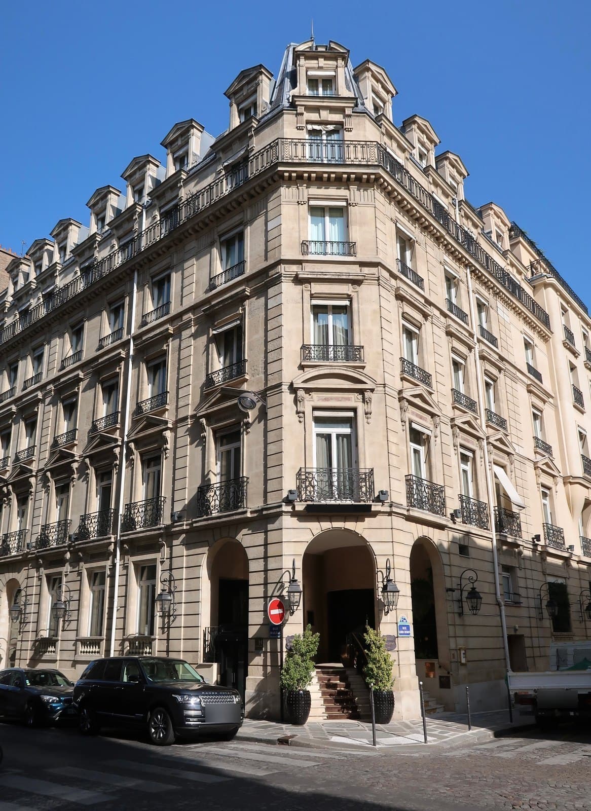Photograph of the cut-stone corner building of the Hôtel Balzac, five storeys with wrought-iron balconies under two levels of mansard dormers and a domed corner pavilion, with an arched entrance up a few steps between two potted topiary trees and lanterns, and parked cars on the street.