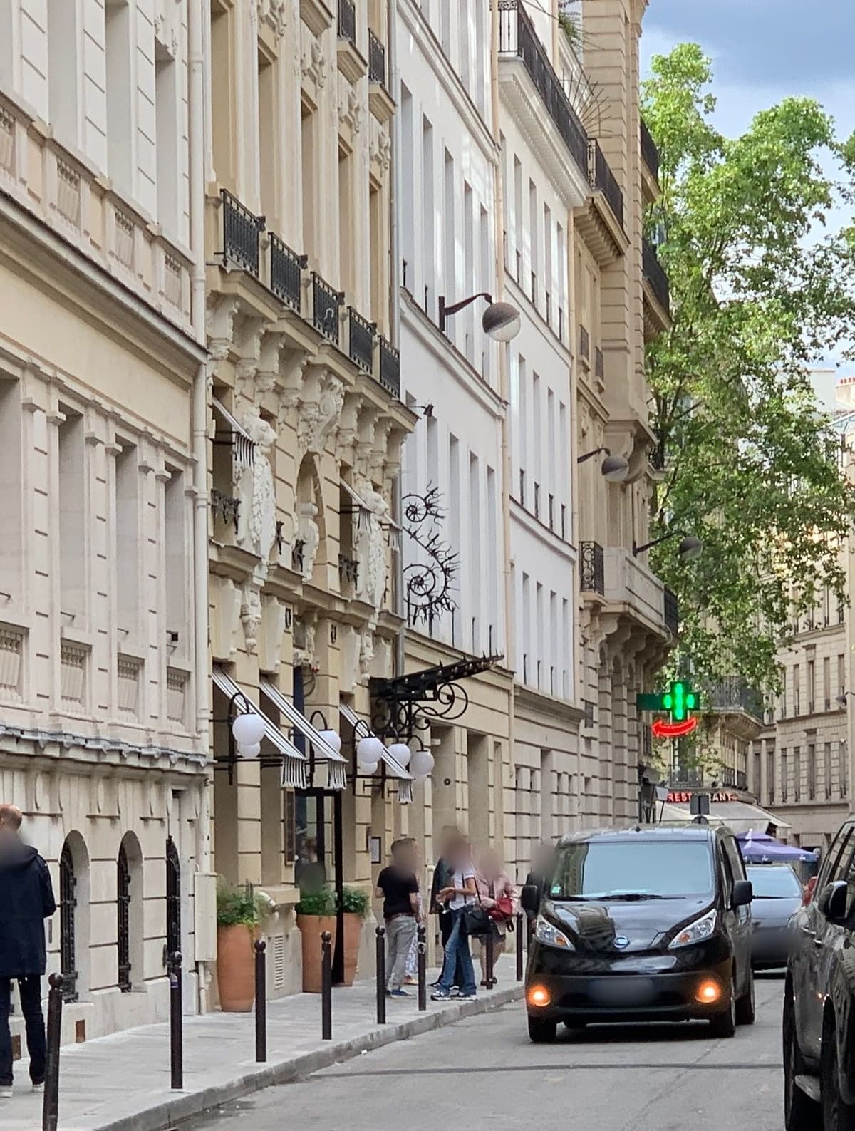 Photograph looking along rue du Helder. On one stone façade, carved figures stand above black-and-white striped awnings, white globe lamps and a wrought-iron sign over the hotel entrance, which is flanked by terracotta planters. A few people stand on the pavement and a black car comes up the street.