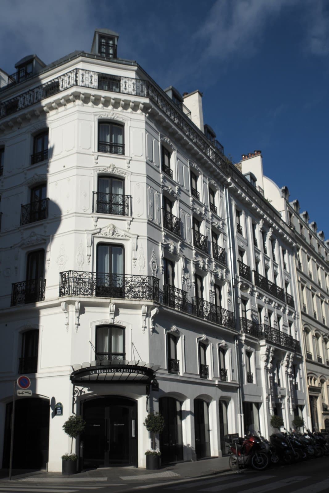 Photograph of the white corner building of Hôtel de Nell on rue du Conservatoire, with wrought-iron balconies, a dark canopy over the corner entrance and scooters parked on the pavement.