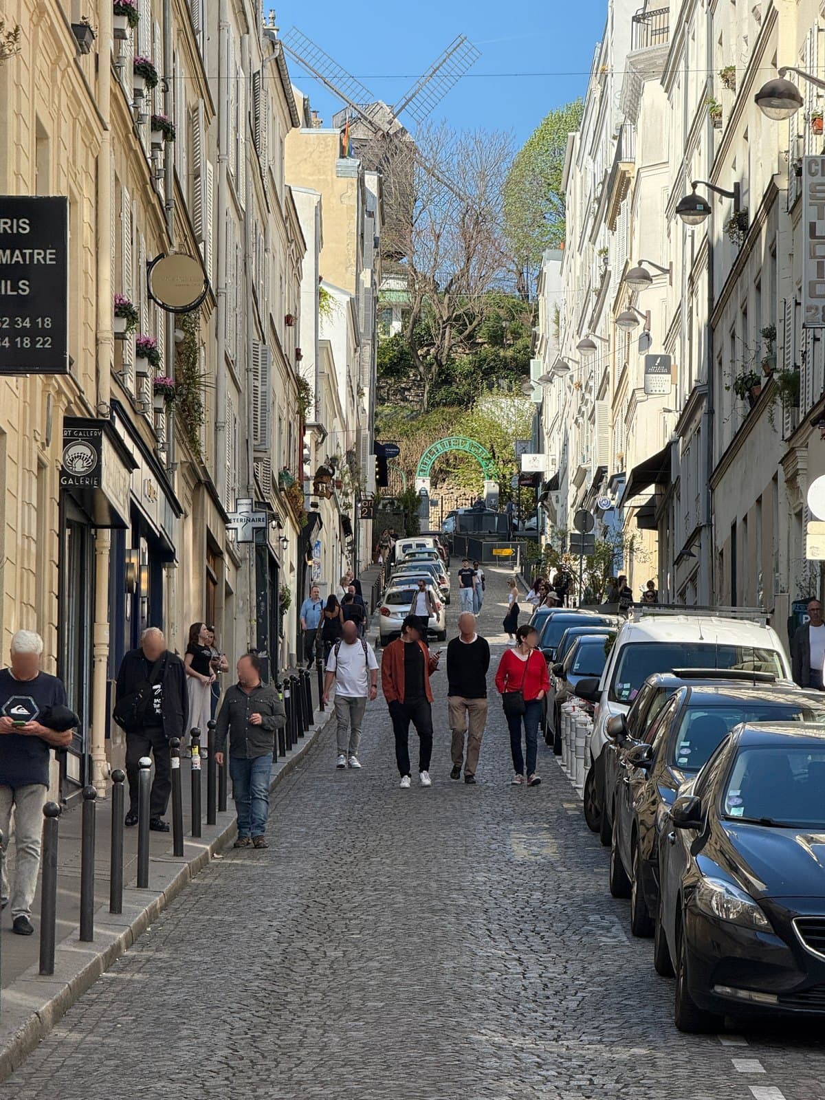 Photograph looking up the steep cobbled rue Tholozé towards a windmill, with cream façades on both sides, a small round hotel sign on the left, parked cars and people walking uphill.
