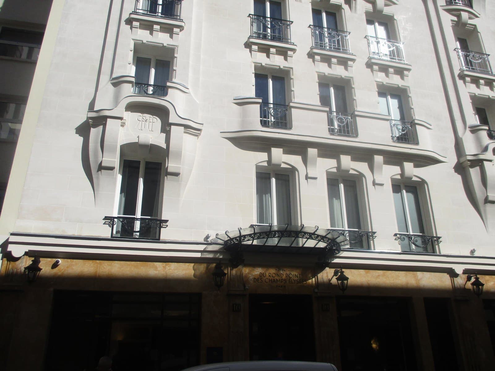 Photograph looking up at the pale stone front of the hotel, with rounded bays on carved brackets, wrought-iron balconies, a monogram carved over a window and a fan-shaped iron-and-glass canopy over the entrance, above a ground floor faced in yellow marble and lettered Hôtel du Rond Point des Champs Élysées.