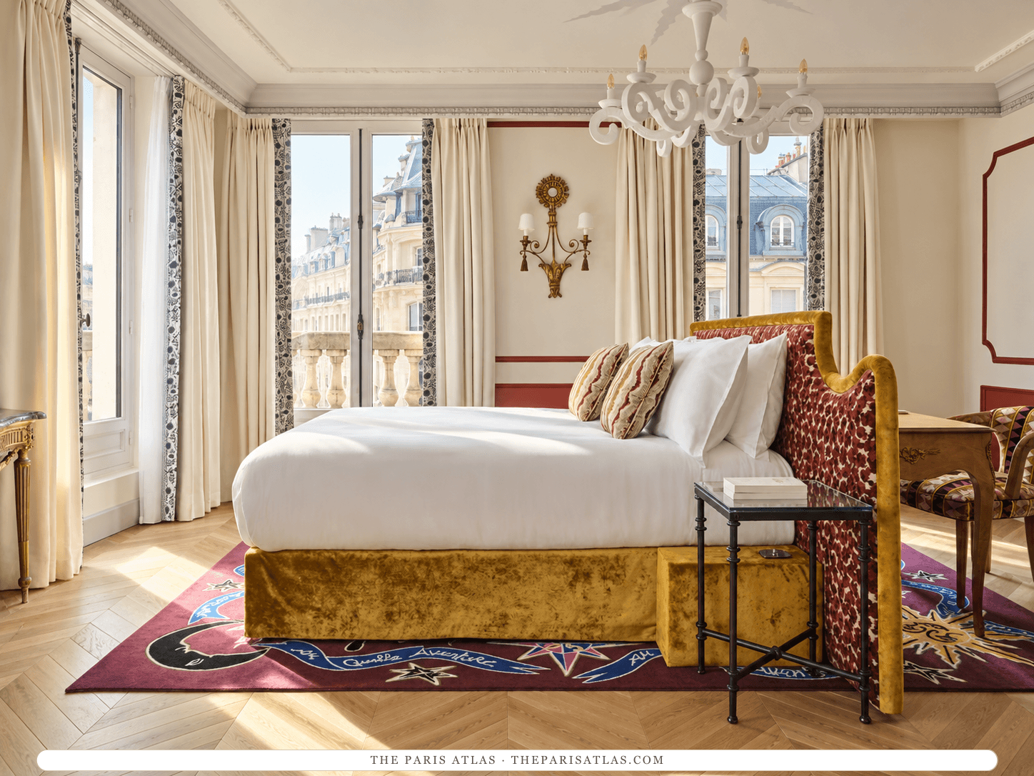 AI-generated illustration of a bedroom at Hôtel L'Aventure: a white bed with a mustard velvet base and a patterned red headboard, a crystal chandelier, red molding, tall French windows and a patterned rug on a herringbone floor.