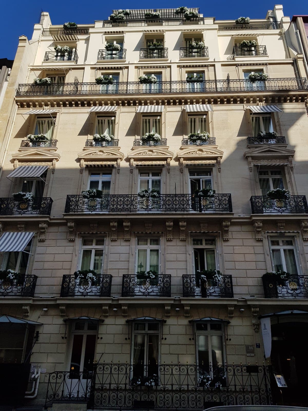 Photograph looking up at the cream stone façade of the Hôtel Lancaster, with wrought-iron balconies, white flowers in window boxes and grey-and-white striped awnings over the windows. A glass canopy covers the entrance on the right at street level.