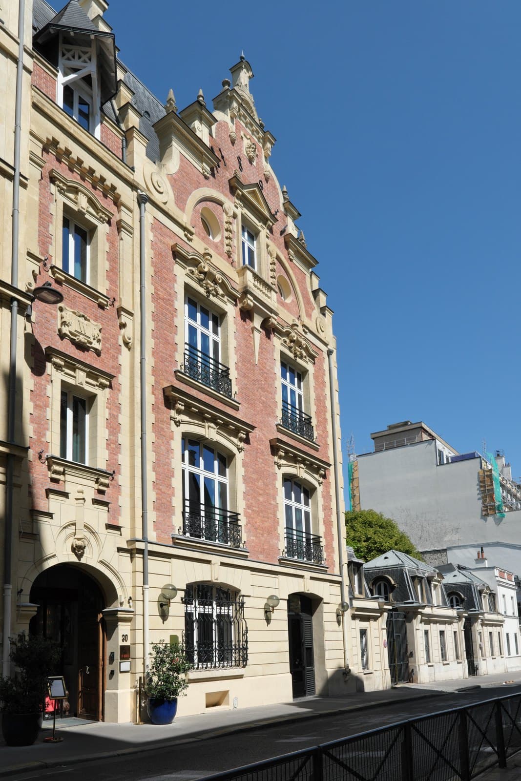 Photograph of the brick-and-stone front of Le Ballu in sunshine, with a stepped Flemish-style gable, a carved W panel, wrought-iron balconies and an arched carriage entrance numbered 30, a menu board and potted plants on the pavement, and lower houses with mansard roofs to the right.