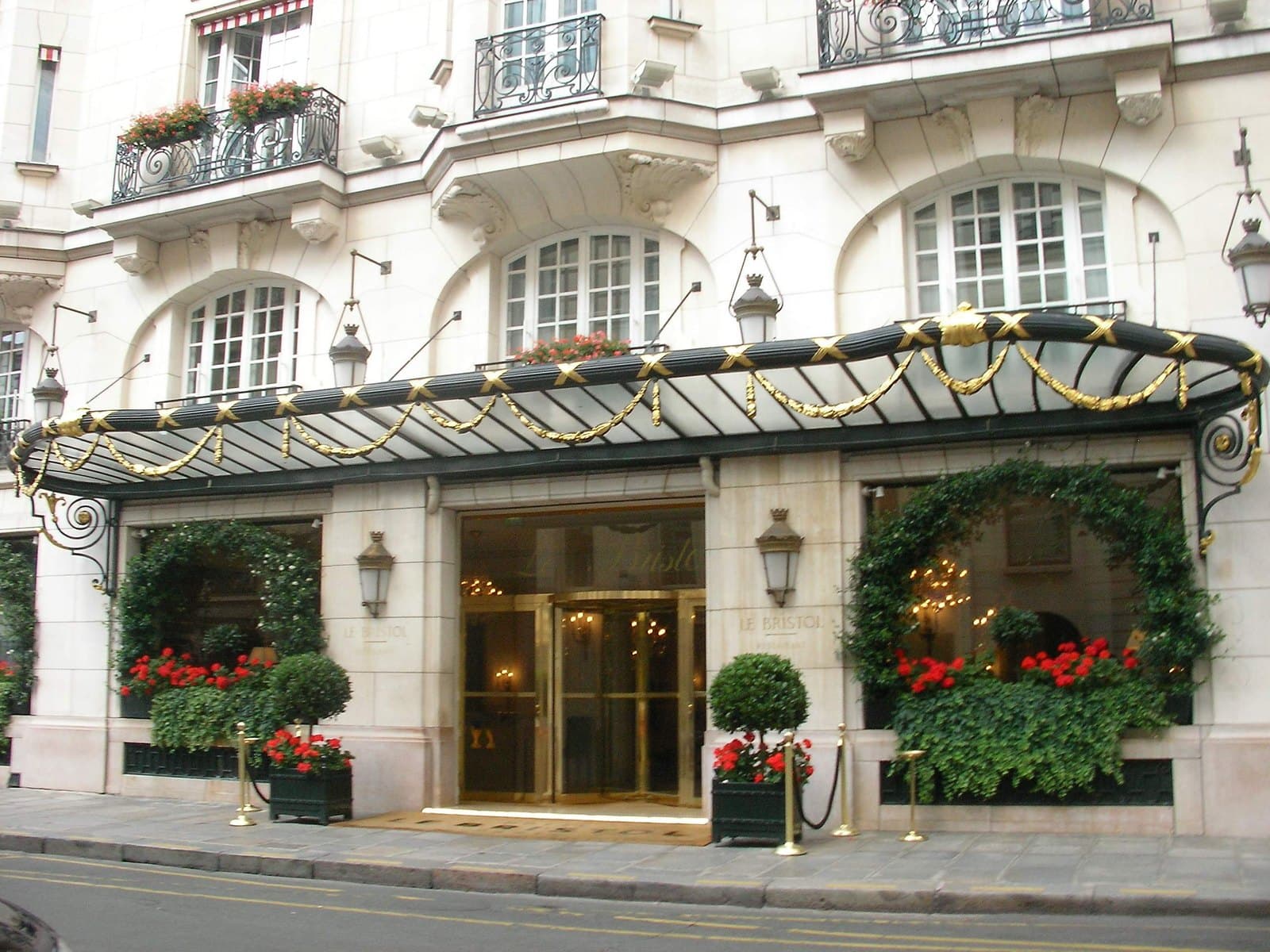 Photograph of Le Bristol's entrance on rue du Faubourg-Saint-Honoré: a pale stone ground floor under a long glass-and-iron canopy hung with gold swags, revolving doors between lanterns, the words Le Bristol carved on the stone, topiary and red geraniums in planters, and windows framed with ivy.