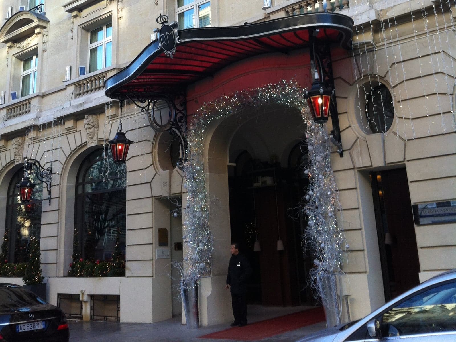 Photograph of the entrance of Le Royal Monceau - Raffles Paris on avenue Hoche: a stone façade with a curved black-and-red canopy topped by an RM monogram, red lanterns, an arched doorway hung with silver Christmas garlands, and a doorman in a dark coat beside a red carpet.