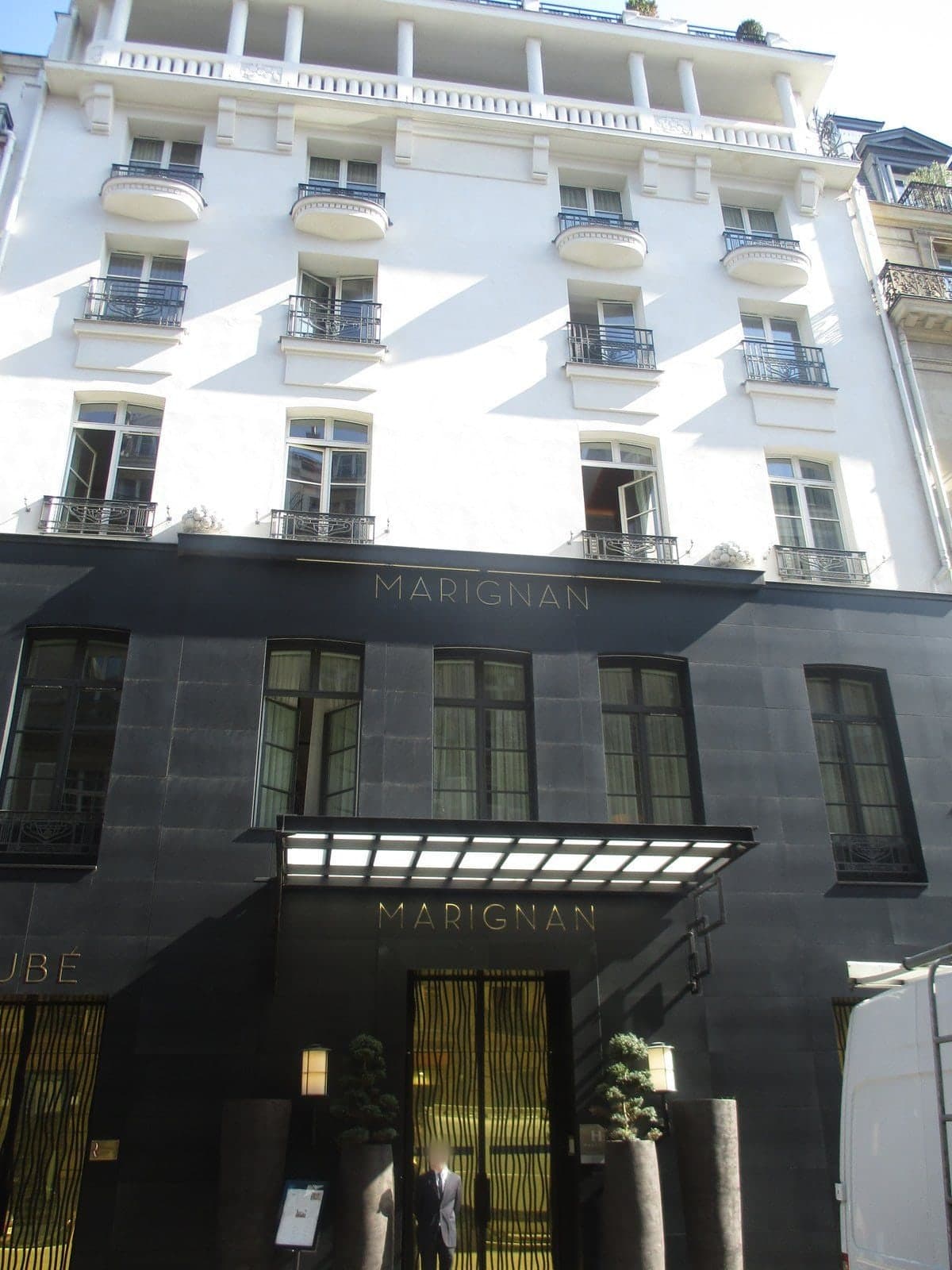Photograph of a white rendered hotel building with rounded balconies and wrought-iron rails above a dark stone ground floor. The word MARIGNAN is lettered across the stone band and above a lit glass canopy over gold entrance doors flanked by potted trees.