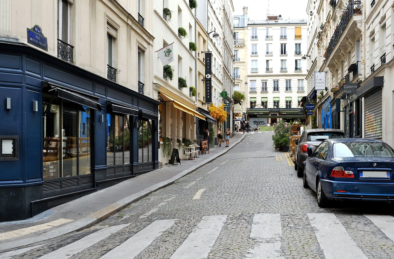 Photograph along cobbled rue Aristide-Bruant, with a cream four-storey building on the left under a yellow scalloped awning, a white flag reading Aristide, window boxes of plants and parked cars.