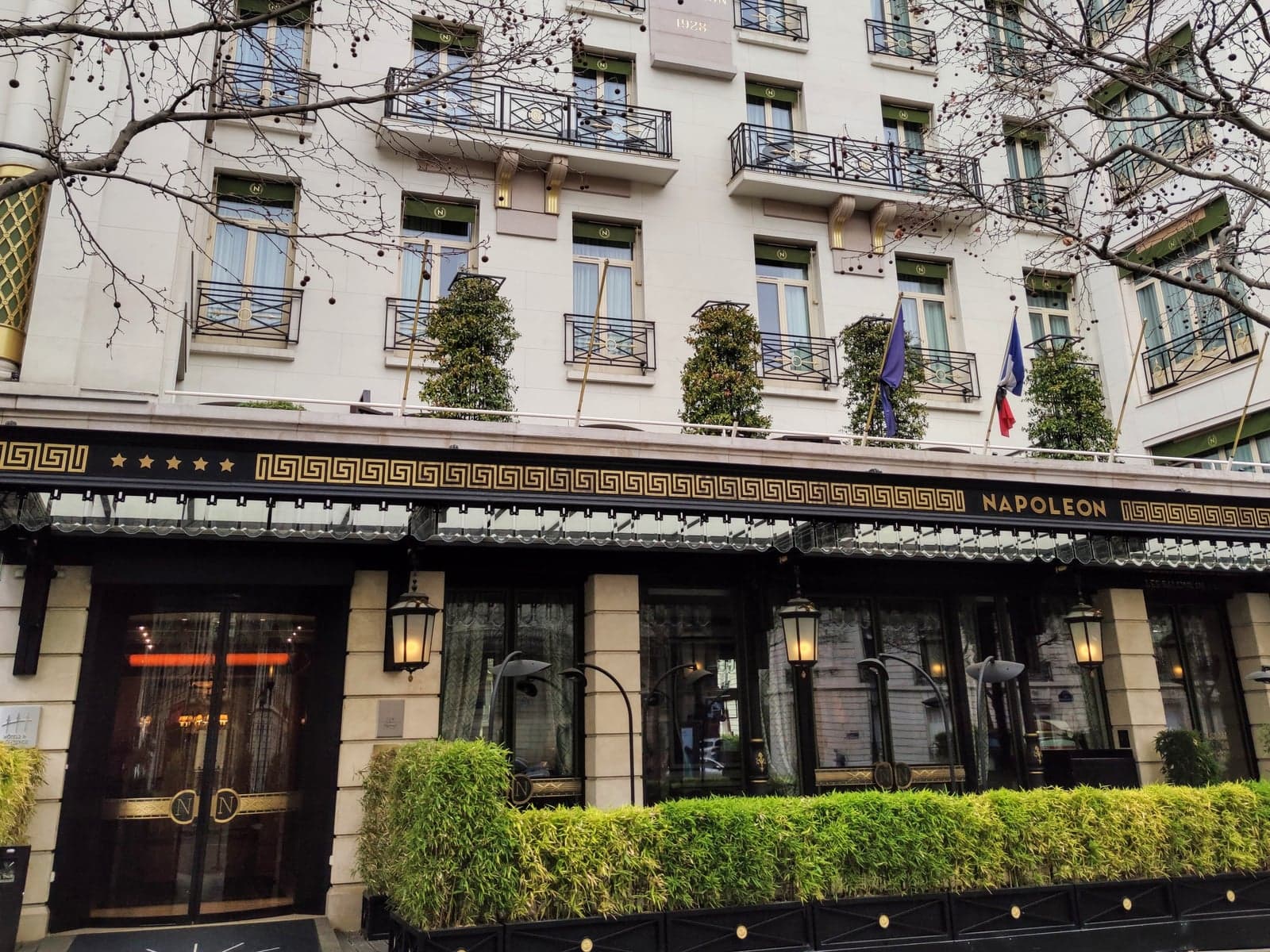 Photograph of Hôtel Napoléon on avenue de Friedland: a white stone façade with green awnings and iron balconies, a black-and-gold canopy reading Napoleon beside five stars, glass entrance doors with N monograms and a low bamboo hedge, seen through bare winter branches.