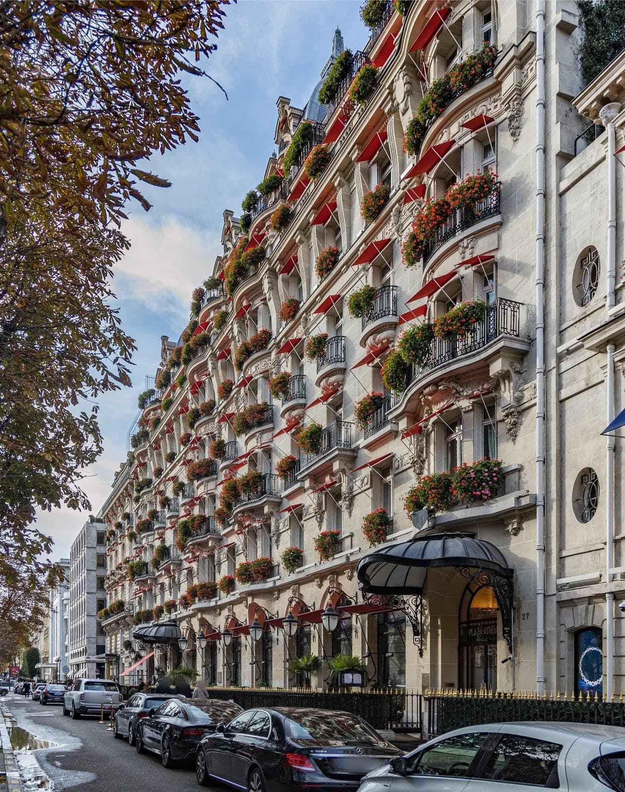 Photograph of the long stone façade of the Hôtel Plaza Athénée on avenue Montaigne in autumn, with red awnings and red geraniums on every balcony, a black glass canopy over the entrance with iron lanterns, and cars parked along the kerb.