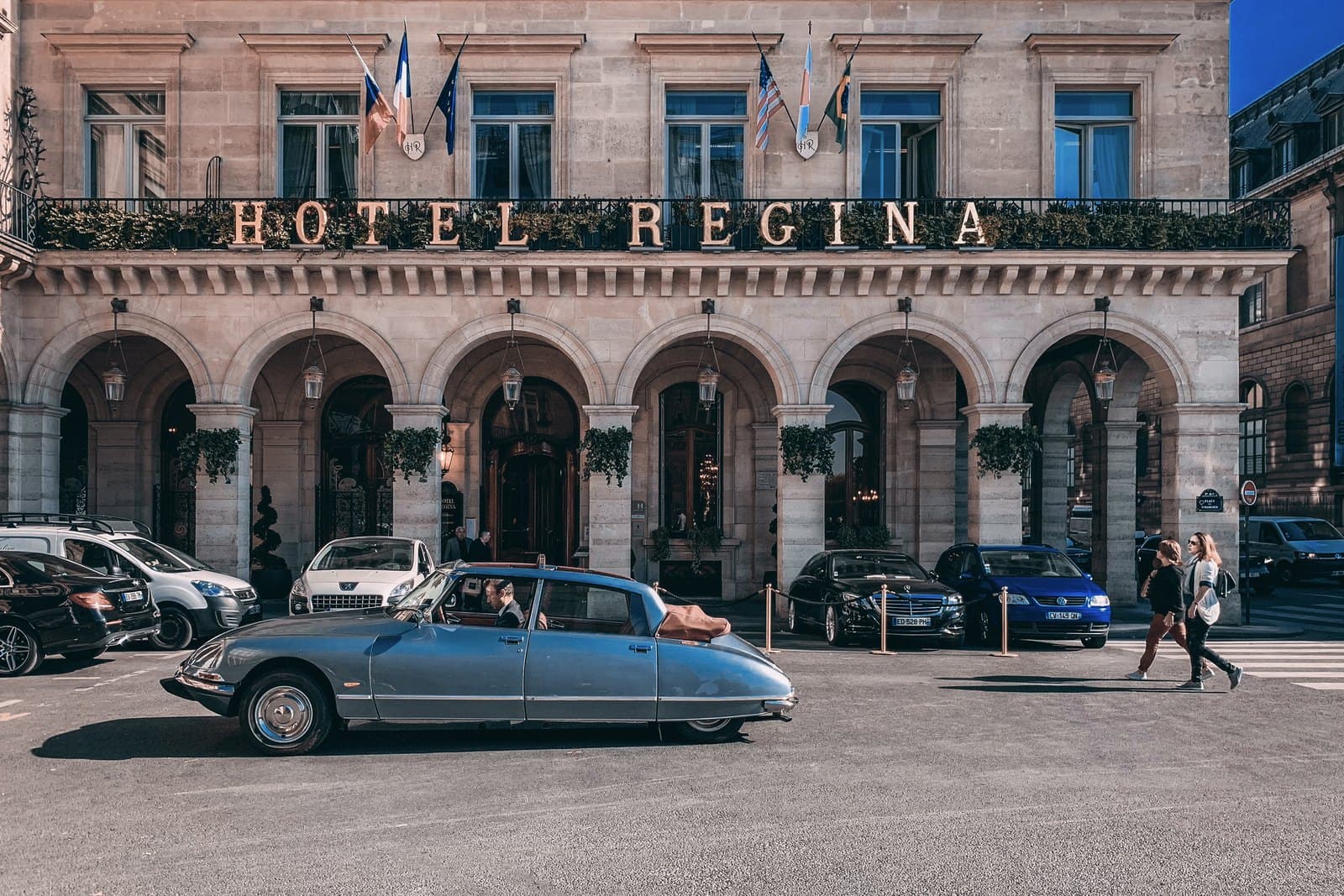 Photograph of Hôtel Regina Louvre's stone front on place des Pyramides: six arches at street level, the words Hotel Regina in gold letters along the first-floor balcony, flags above, and a pale blue vintage Citroën DS driving past.